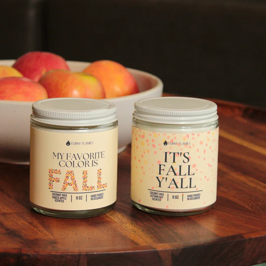 Two 9 oz Funny Flames fall scented candles labeled Baked Apple and Autumn Day on wooden table with apples in background