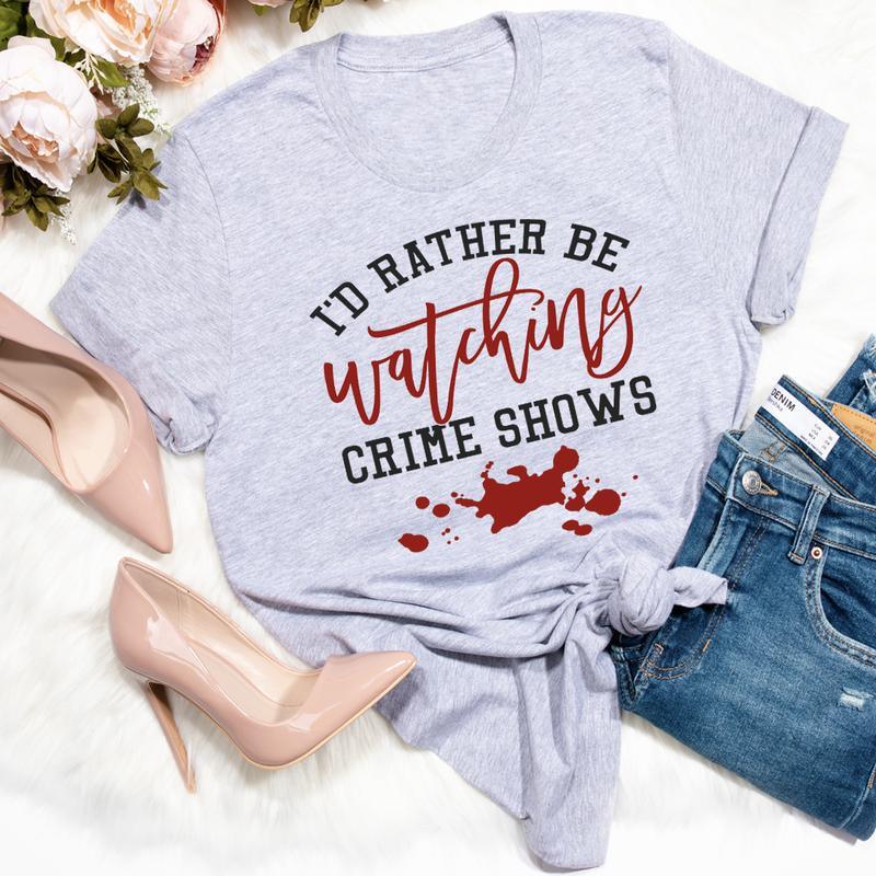 Light gray t-shirt with text I'd rather be watching crime shows and a red blood splatter design, paired with blue jeans and beige high heels