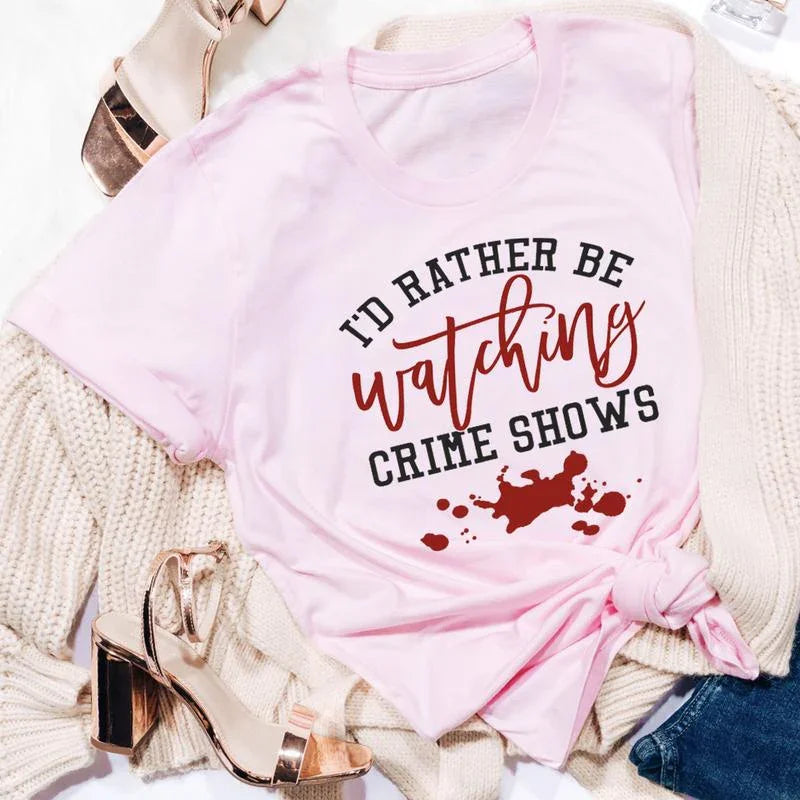 Pink t-shirt with black and red text 'I'd rather be watching crime shows' and blood splatter design, styled with beige knitwear and rose gold heels