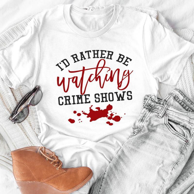 White t-shirt with text 'I'd rather be watching crime shows' and red blood splatter graphic, styled with jeans, brown boots, and sunglasses