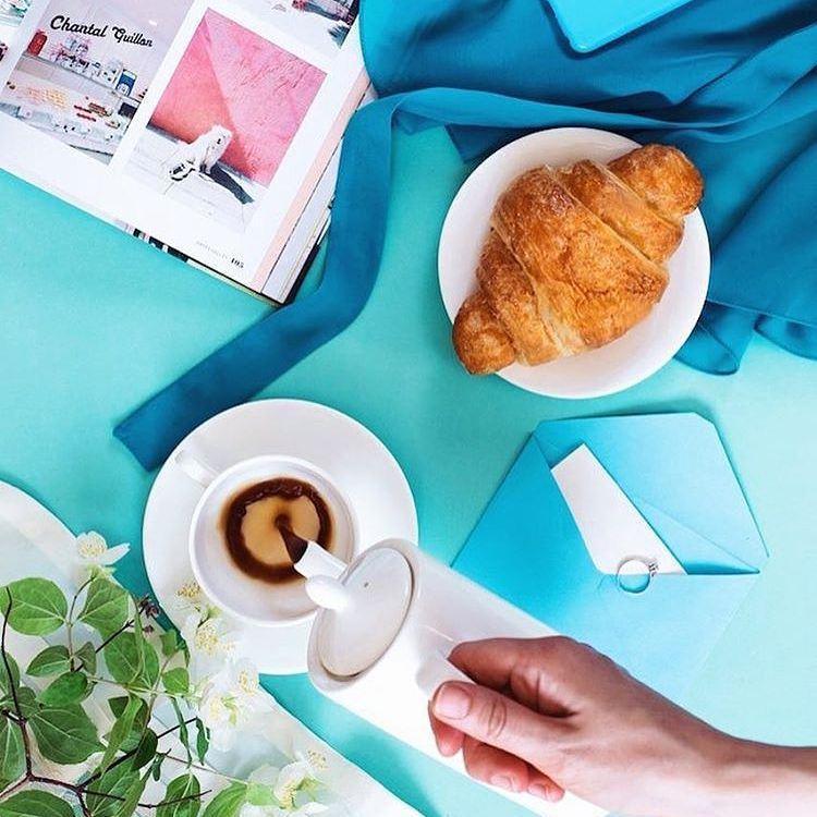 Flat lay with croissant, coffee, magazine, and flowers on a bright turquoise background.