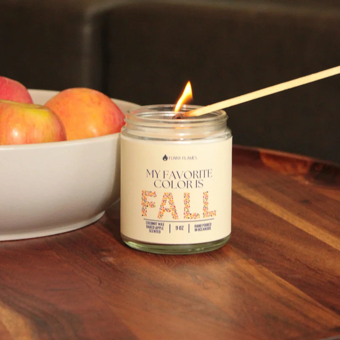 Lit fall-scented candle reading 'My Favorite Color is Fall' next to a bowl of apples on wooden table