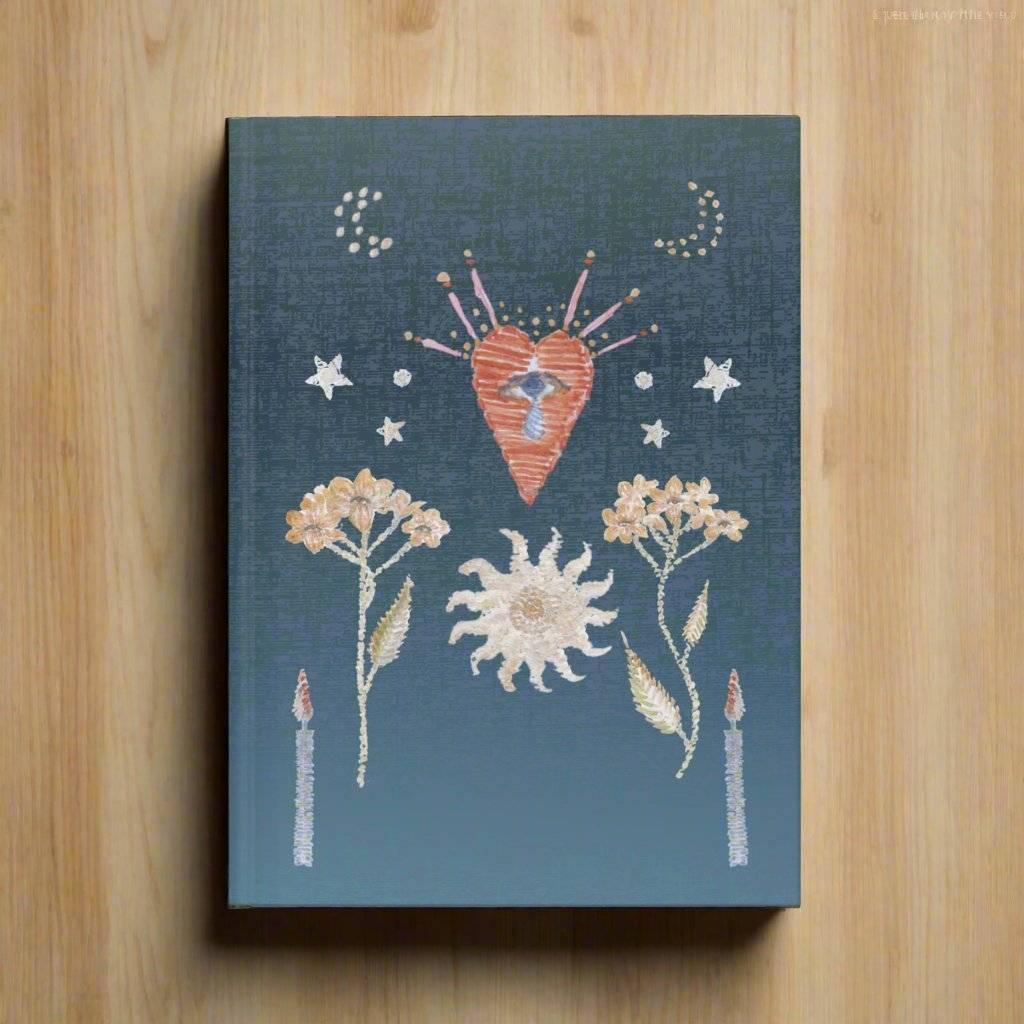 Blue notebook with embroidered heart, flowers, sun, stars, crescent moons, and candles on wooden surface