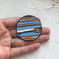 Hand holding round enamel pin with colorful cosmic planet design, Qu!rk Boutique gift accessory