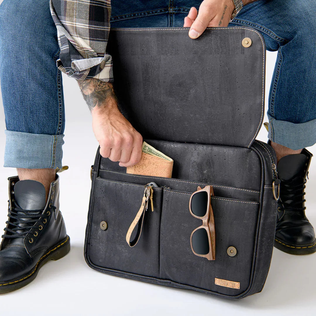 Man with rolled-up jeans and black boots holding cork wallet and sunglasses near black sustainable bag