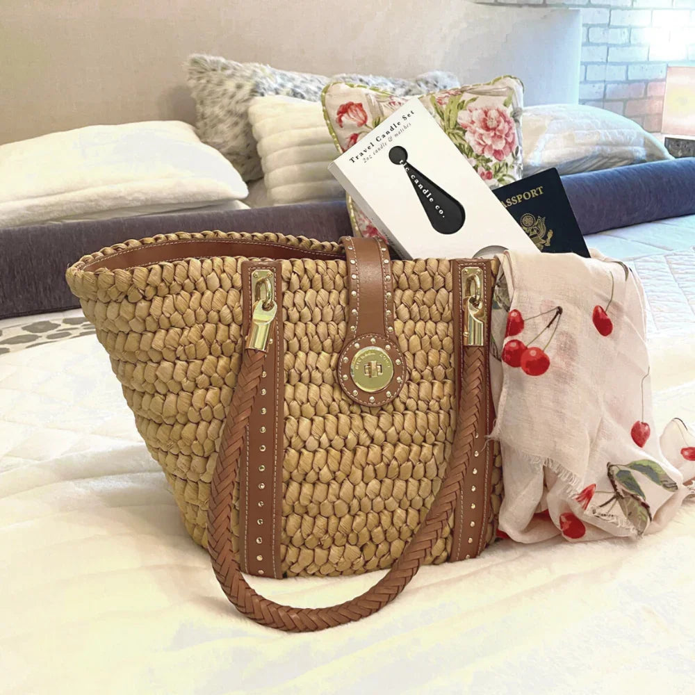 woven straw tote bag with leather straps on bed, holding travel candle set, cherry-print scarf, and passport