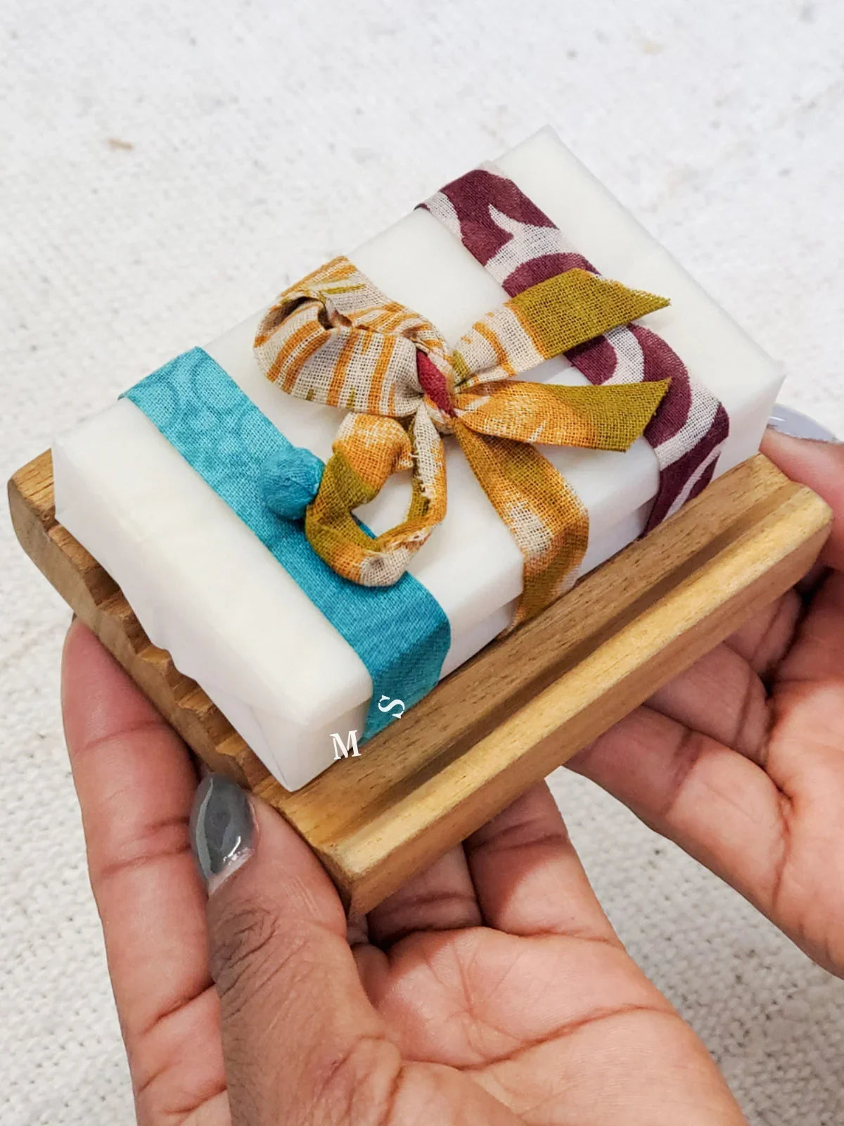 Hands holding white artisan soap bar with colorful fabric ribbons on wooden soap dish