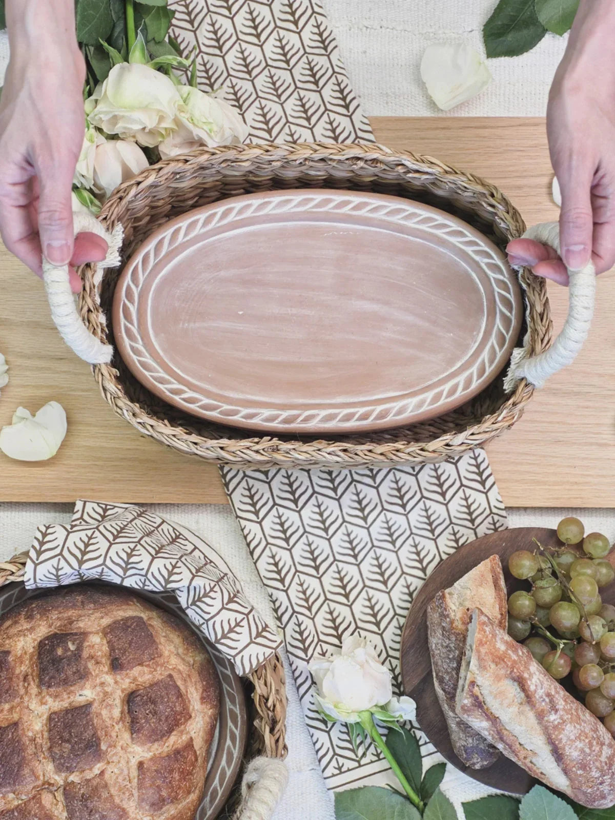 Hands holding woven wicker tray with ceramic oval plate, bread, grapes, and white flowers on patterned cloth