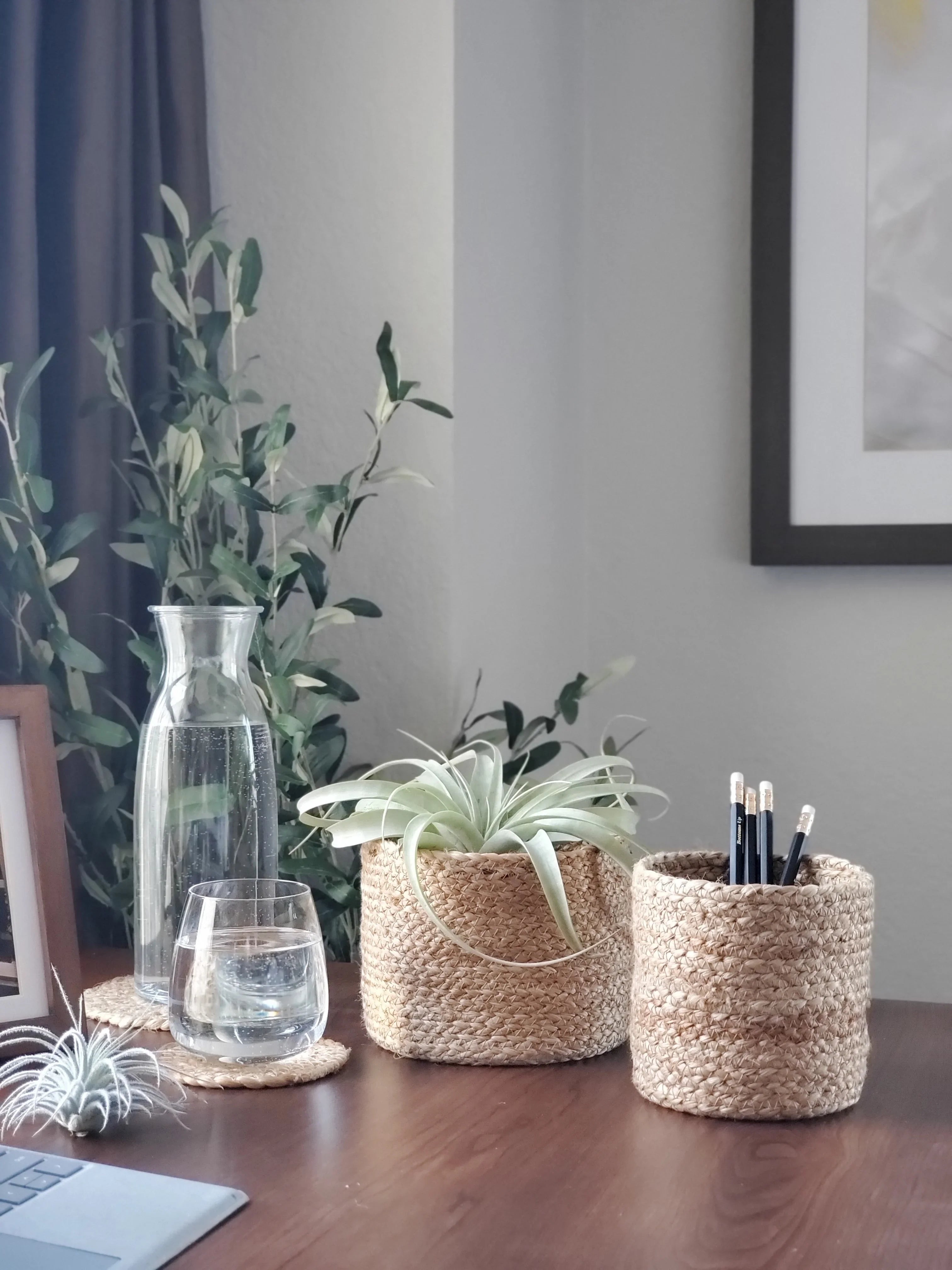 Minimalist desk setup with woven baskets holding plants and pencils, glass carafe and glass of water