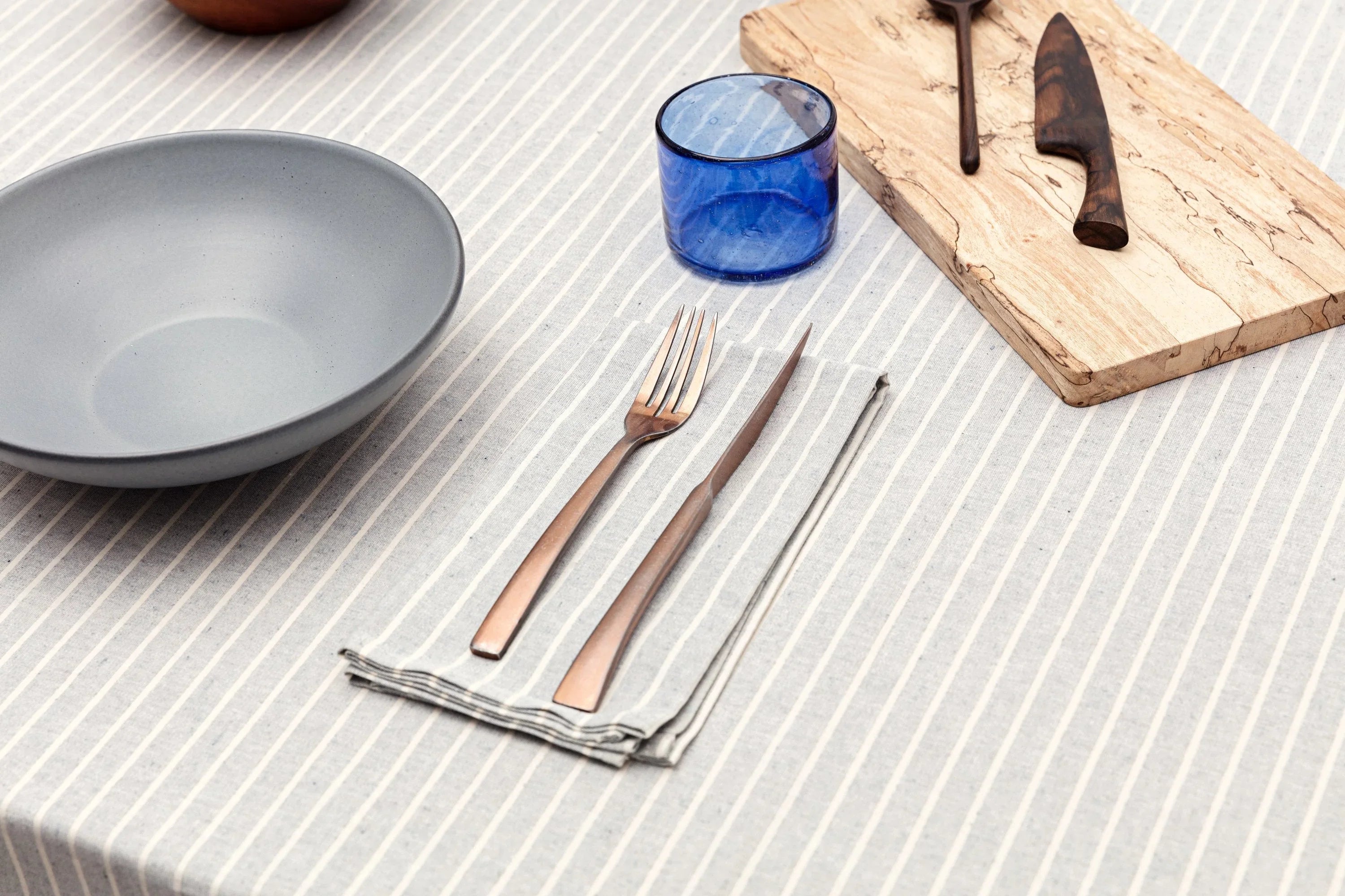 Modern table setting with blue bowl, striped napkin, bronze cutlery, blue glass, wood board at Qu!rk Boutique