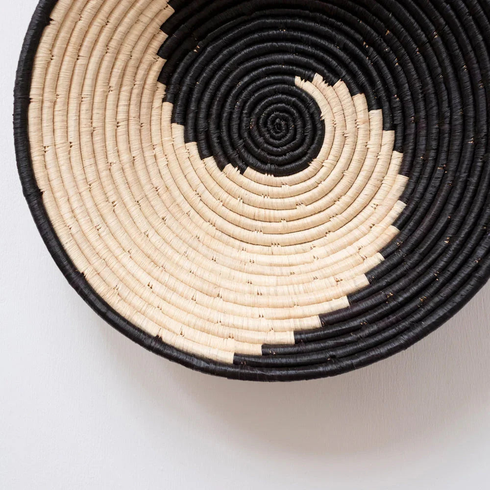 Handwoven black and natural spiral basket on white wall, sustainable artisan home decor.