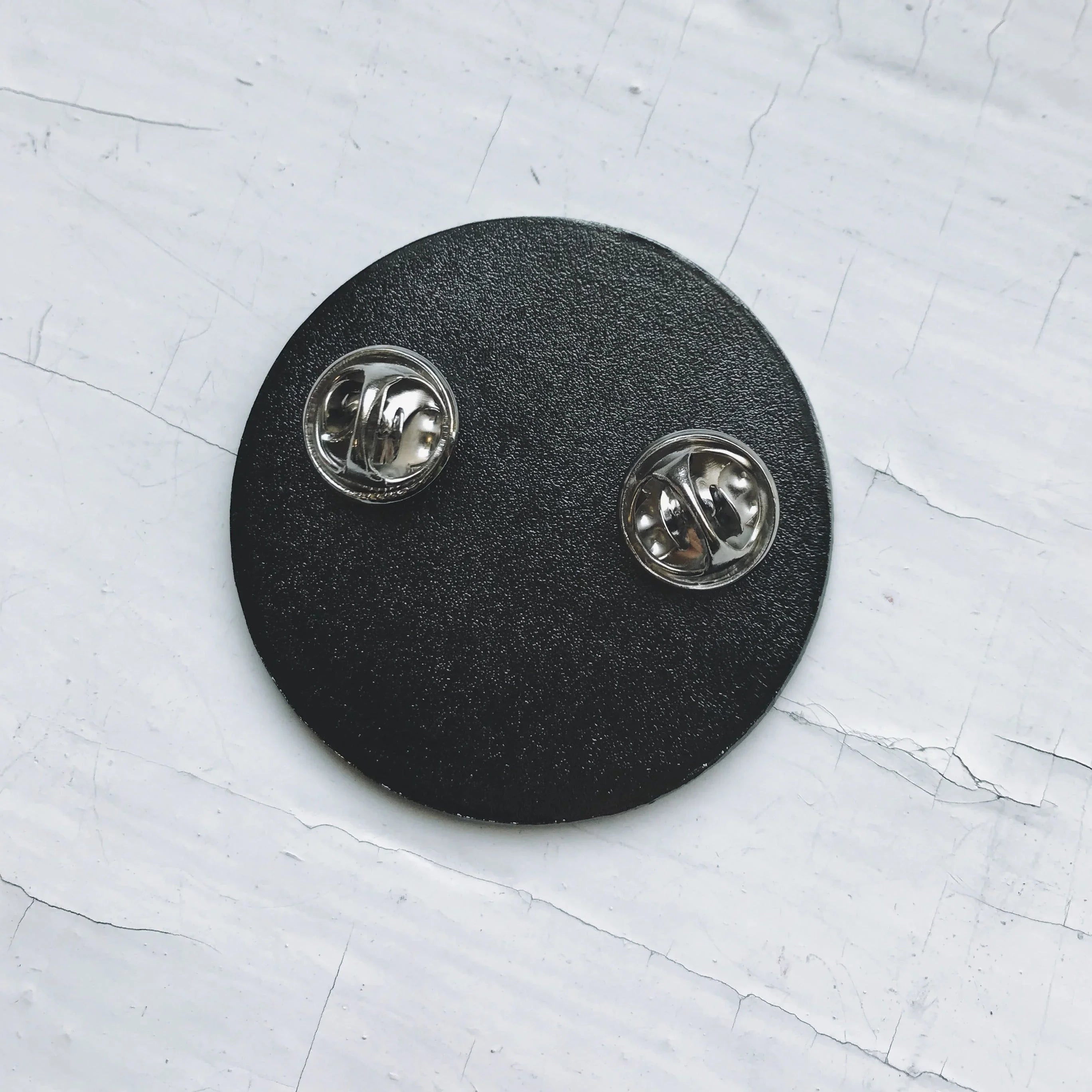 Black round pin backing with two silver butterfly clutch fasteners on white cracked surface