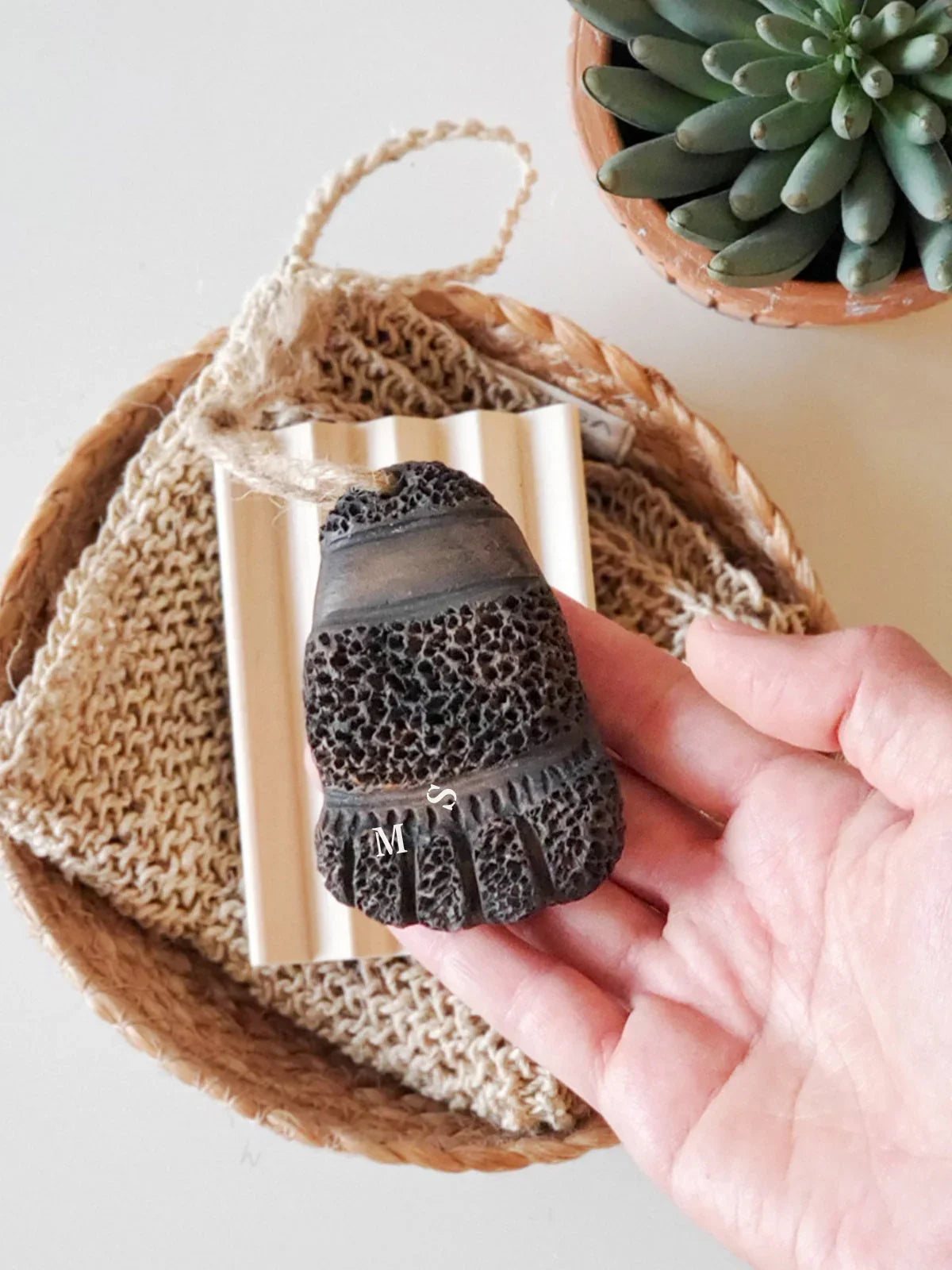 Hand holding natural pumice stone with rope, knitted cloth in basket, and potted succulent plant