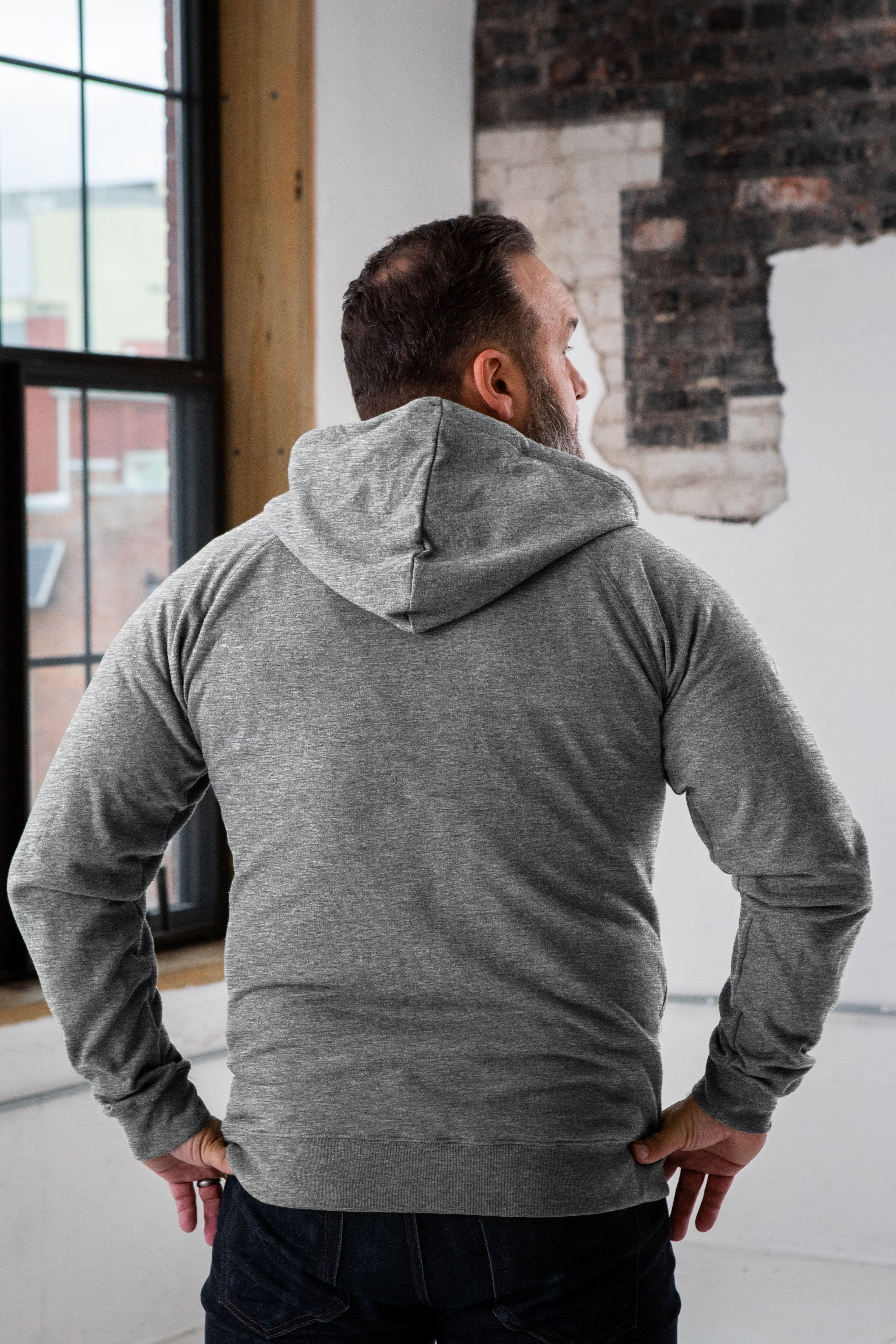 Man in grey hoodie from Qu!rk Boutique stands indoors by loft window with exposed brick wall