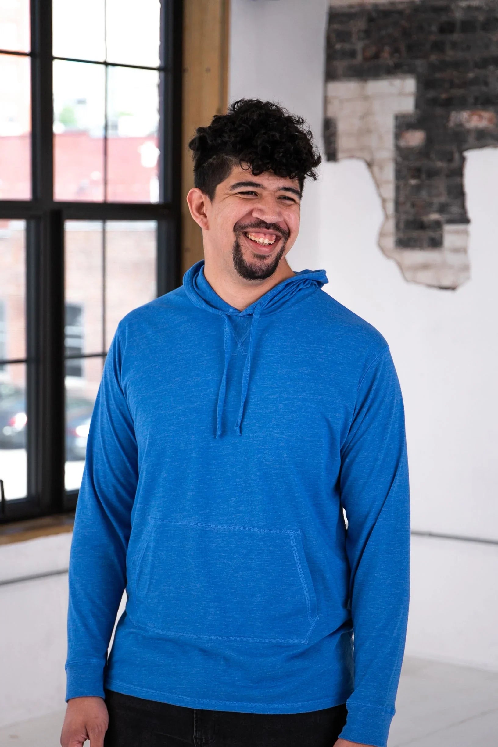 Man smiling in bright blue hoodie, casual fashion at Qu!rk Boutique with urban window backdrop