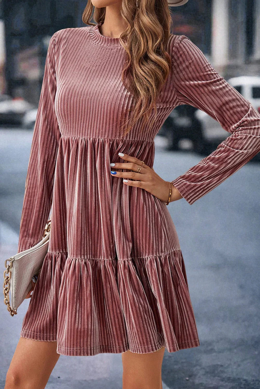 Woman in a mauve velvet long-sleeve dress, accessorized with a white clutch, outdoor setting