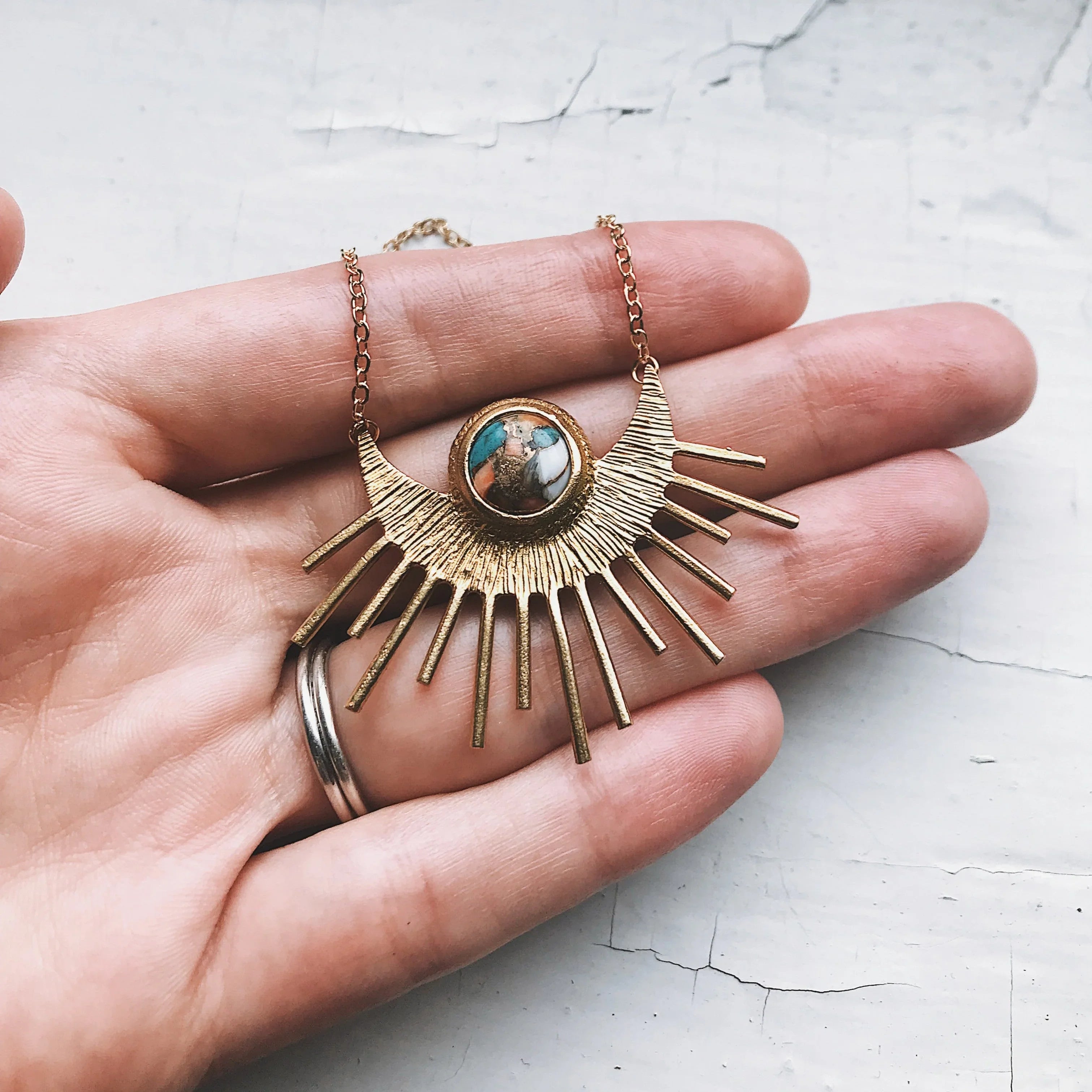 Hand holding gold sunburst necklace with turquoise and white stone pendant on cracked white surface