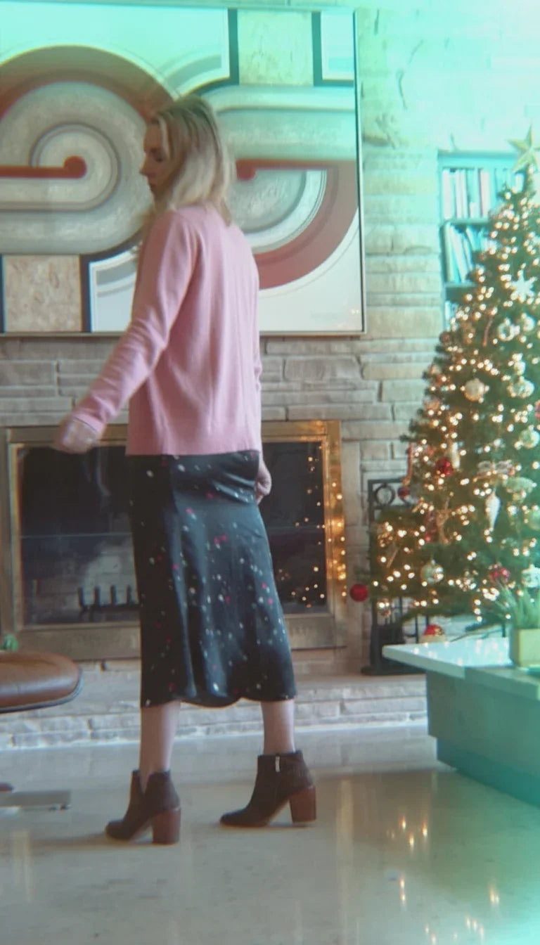 Woman in pink sweater and black floral skirt standing by decorated Christmas tree near fireplace