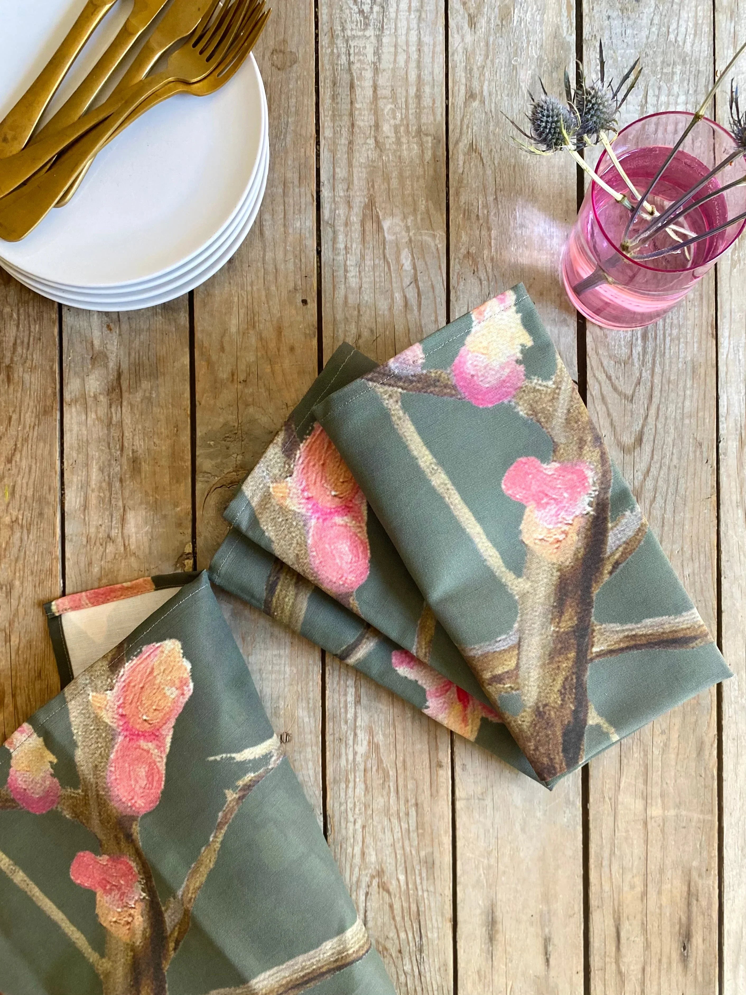 Folded floral print napkins on rustic wooden table with gold flatware on white plates and pink vase with thistles