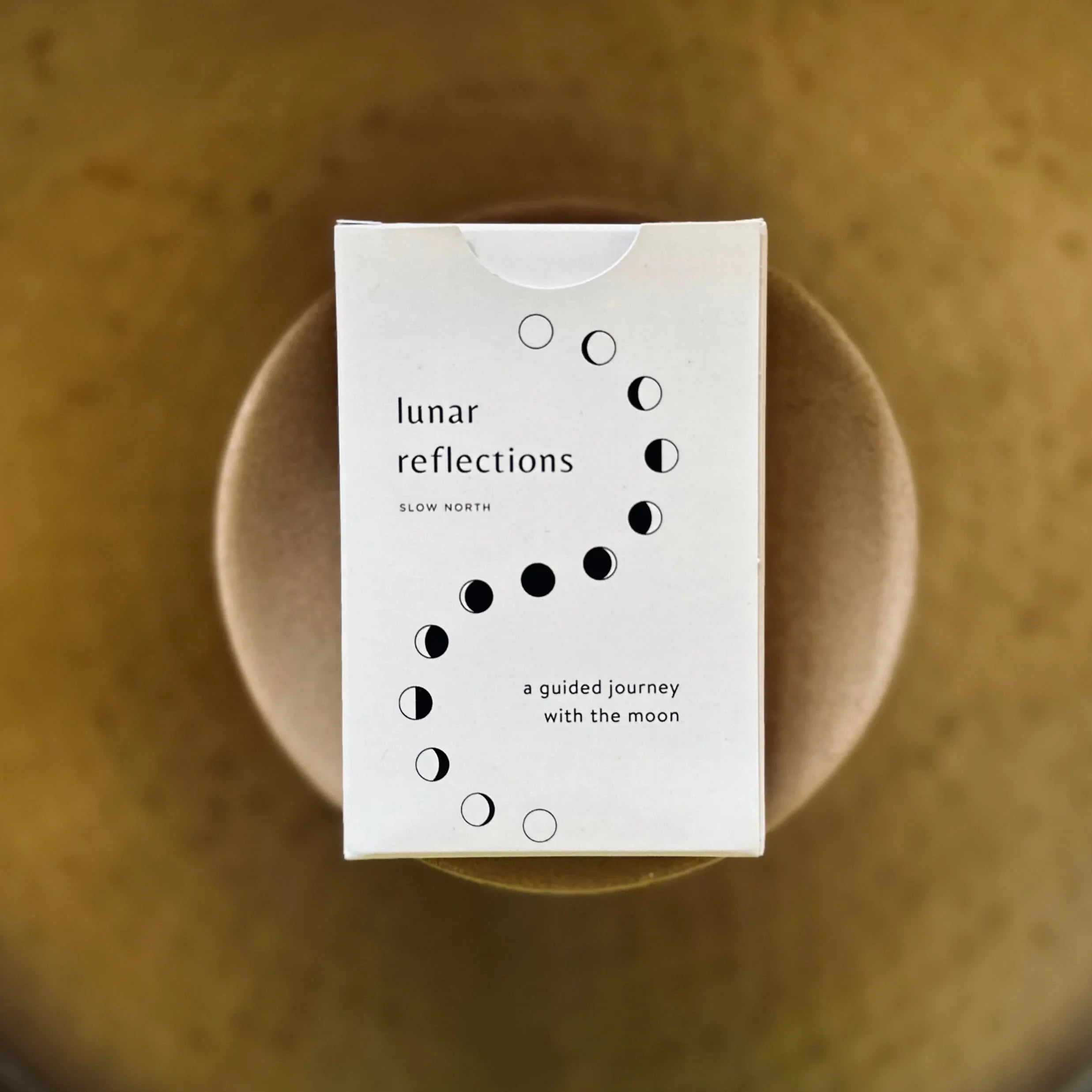 White box labeled lunar reflections by Slow North with moon phase illustrations, placed on a round brown surface