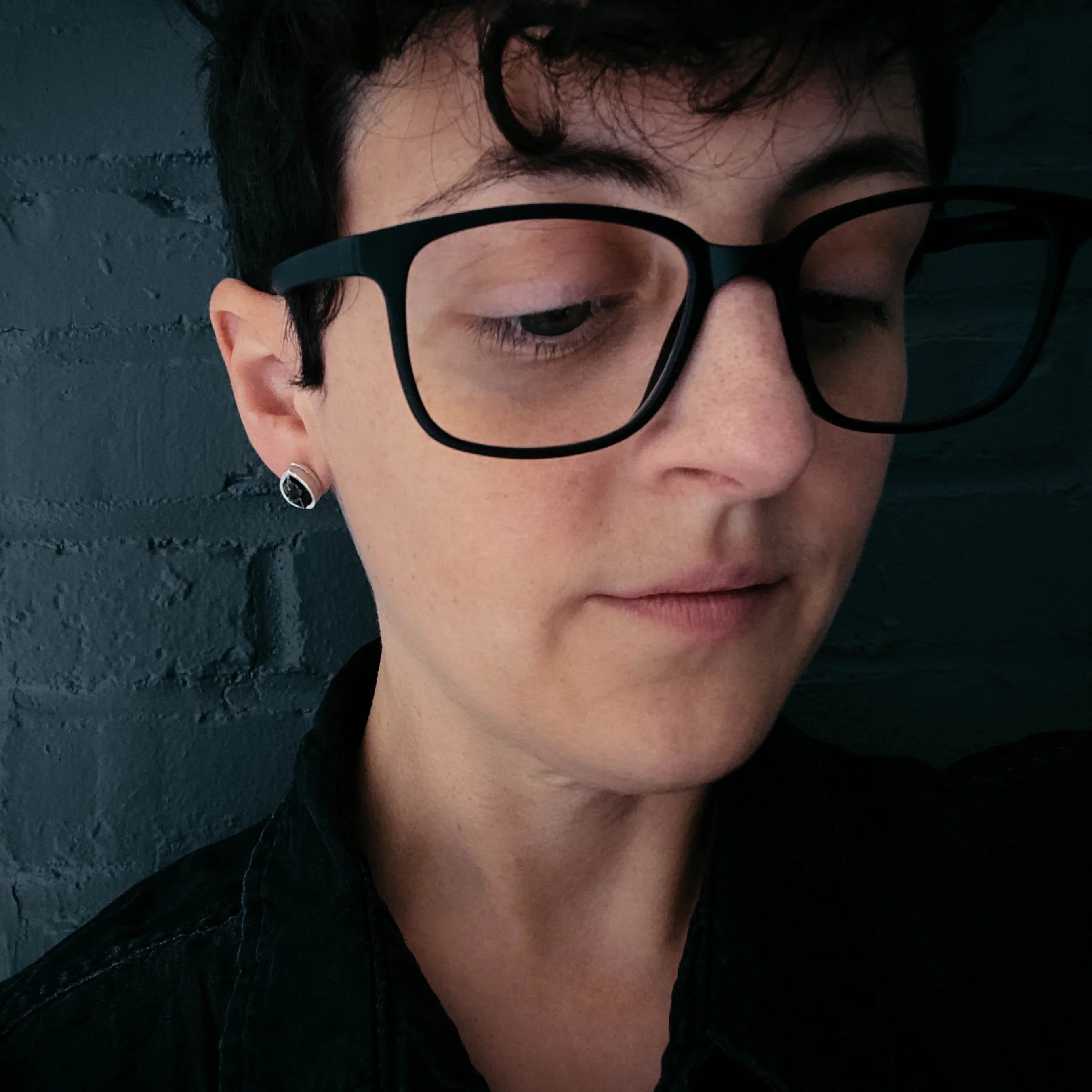 Person with short hair, black glasses, and minimalist earrings near dark brick wall, Qu!rk Boutique style