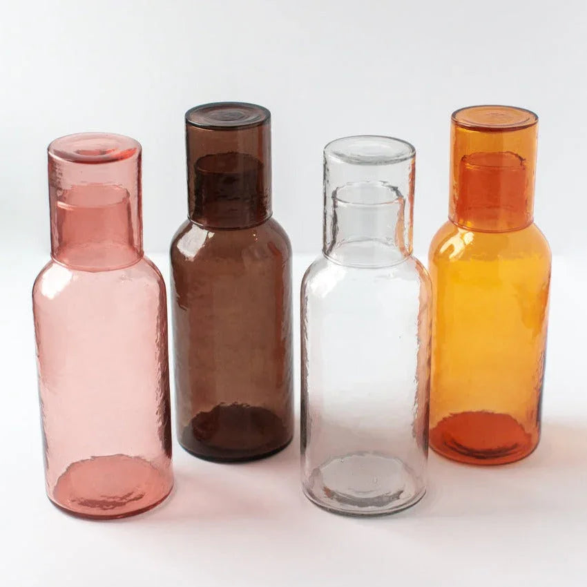 Set of four colorful recycled glass water carafes in pink, brown, clear, and amber, on white background
