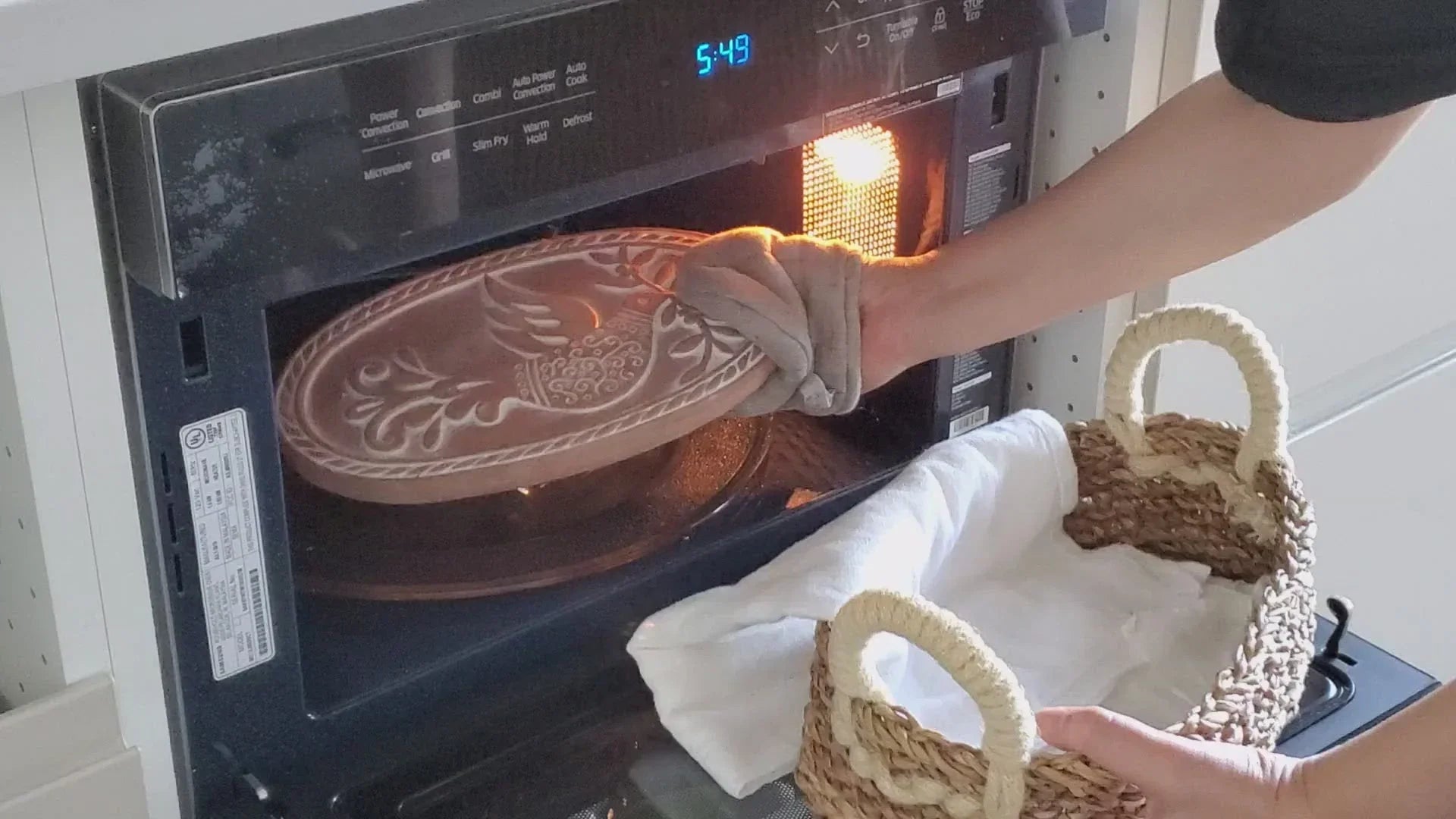 Person using oven mitt to remove decorative brown baking dish from microwave oven near wicker basket with white cloth