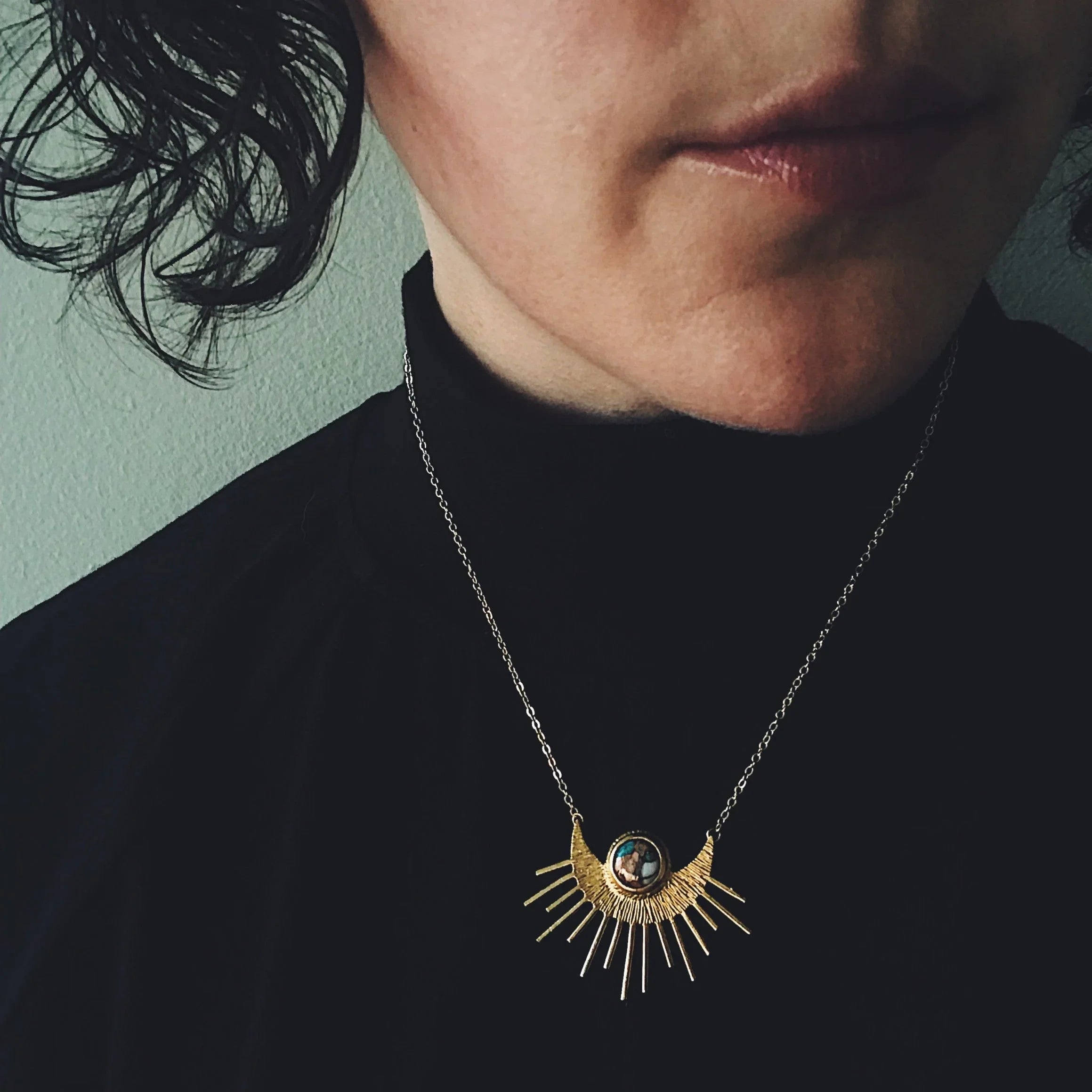Person wearing a gold sunburst necklace with a turquoise stone pendant on a silver chain and black turtleneck