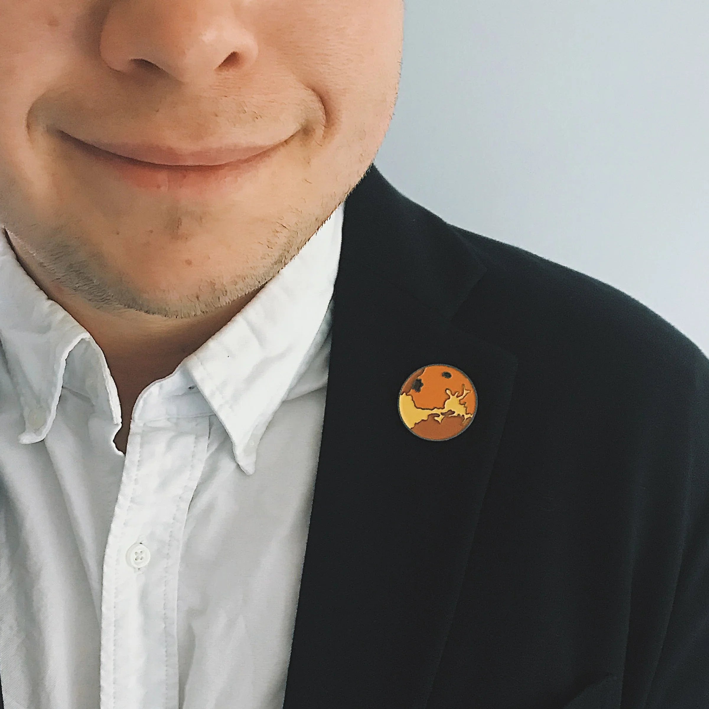 Close-up of a man wearing a white shirt and black blazer with an orange and yellow enamel pin on the lapel