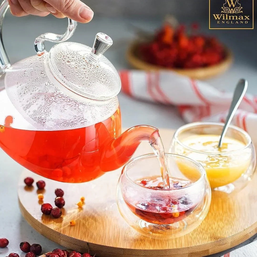 Hand pouring herbal tea from glass teapot into double-walled cup, with honey, berries, and Wilmax England branding visible, on a cozy wooden tray. Quirk Boutique gift shop style.