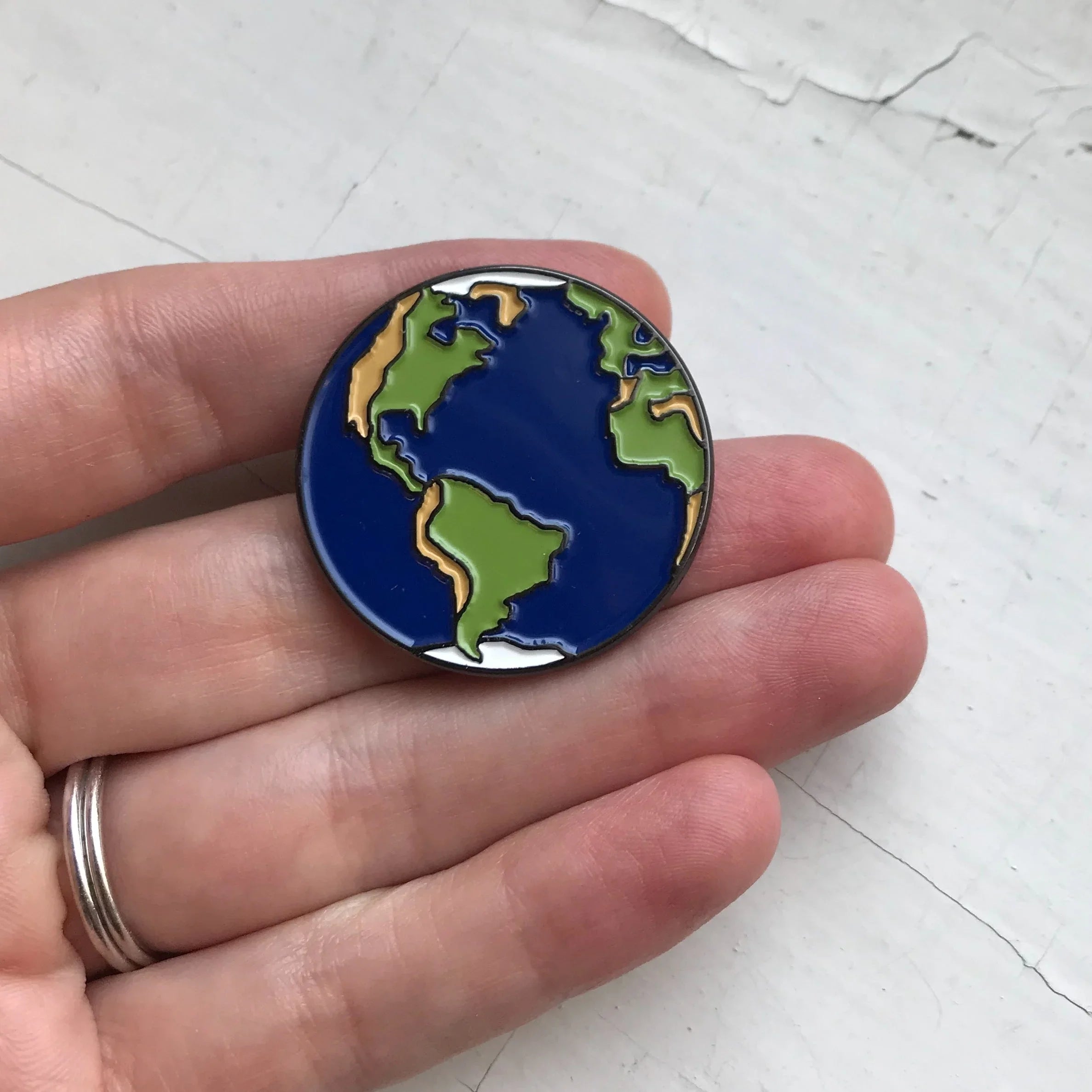 Hand holding a small enamel pin shaped like Earth with blue oceans and green continents