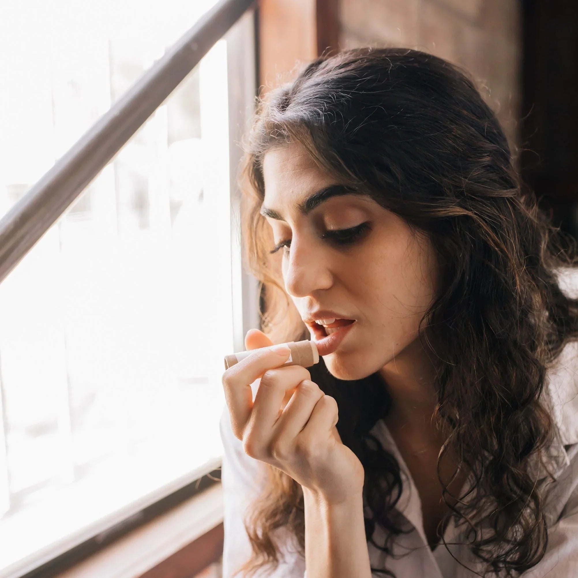 Woman applying natural lip balm by window, eco-friendly beauty at Qu!rk Boutique