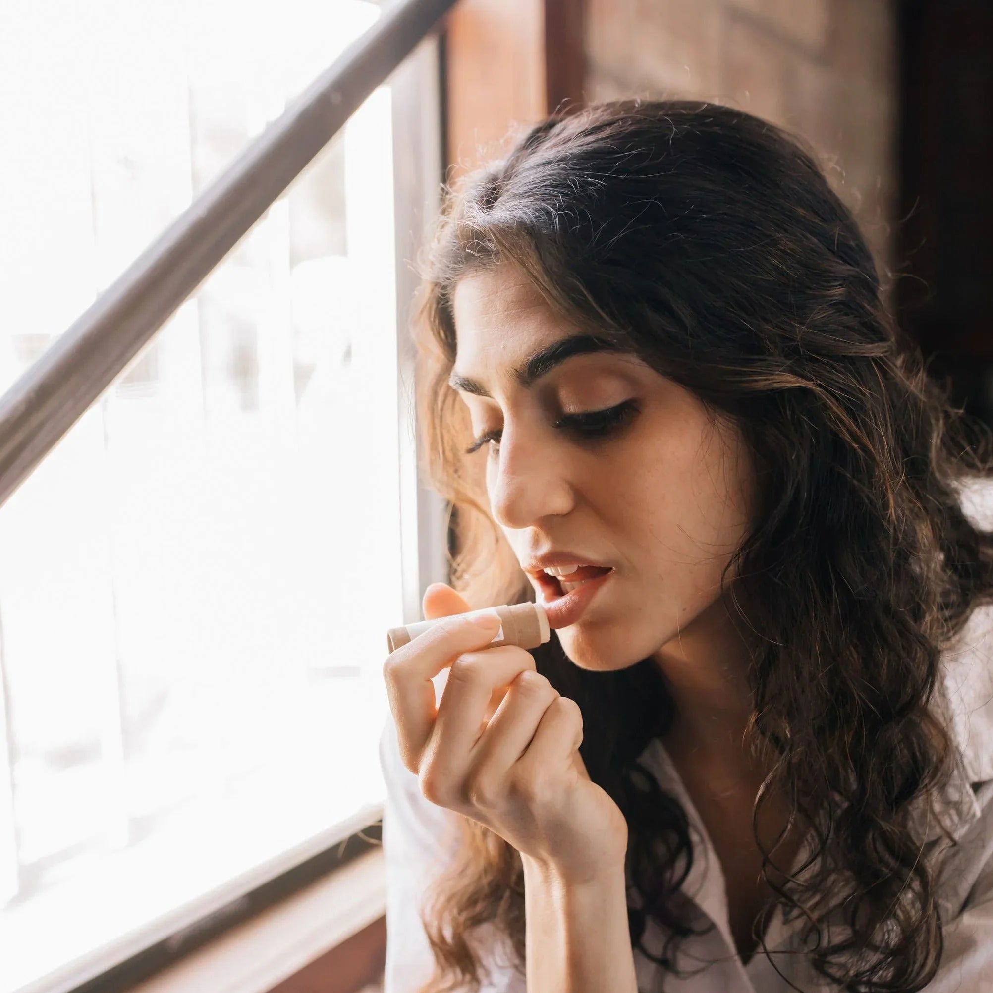 Woman applying natural lip balm by window, eco-friendly beauty at Qu!rk Boutique