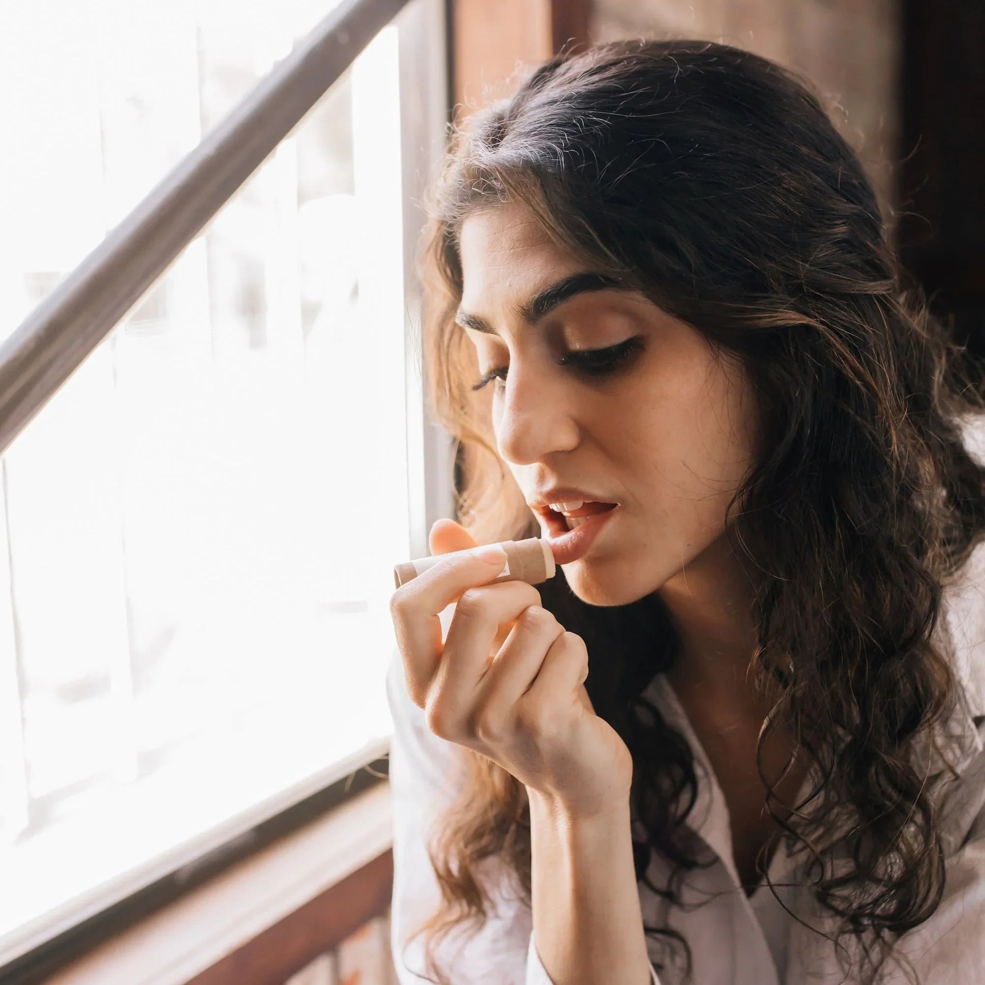 Woman applying natural lip balm by a window, Qu!rk Boutique eco-friendly beauty product