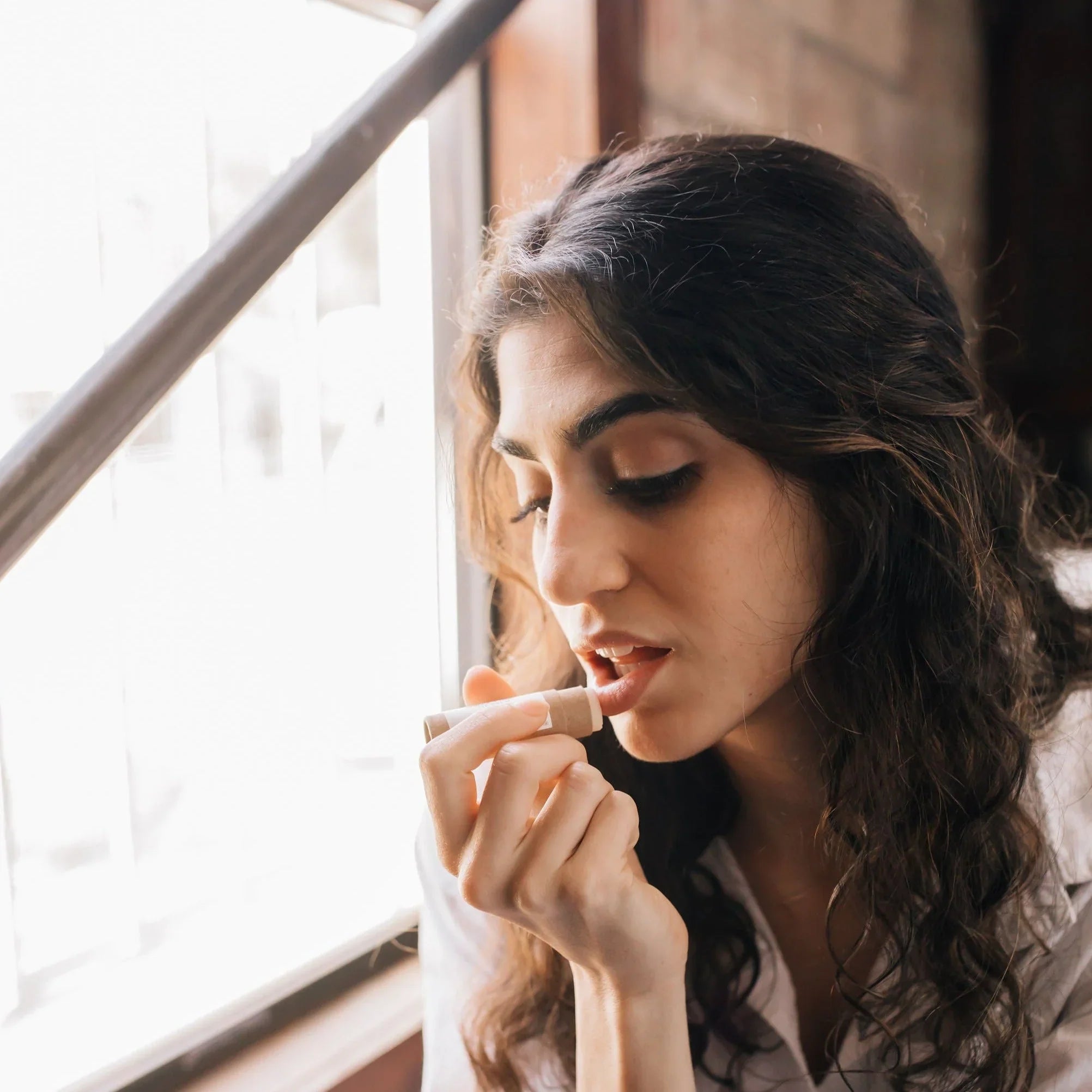 Young woman with dark hair applying natural lip balm near a sunlit window indoors