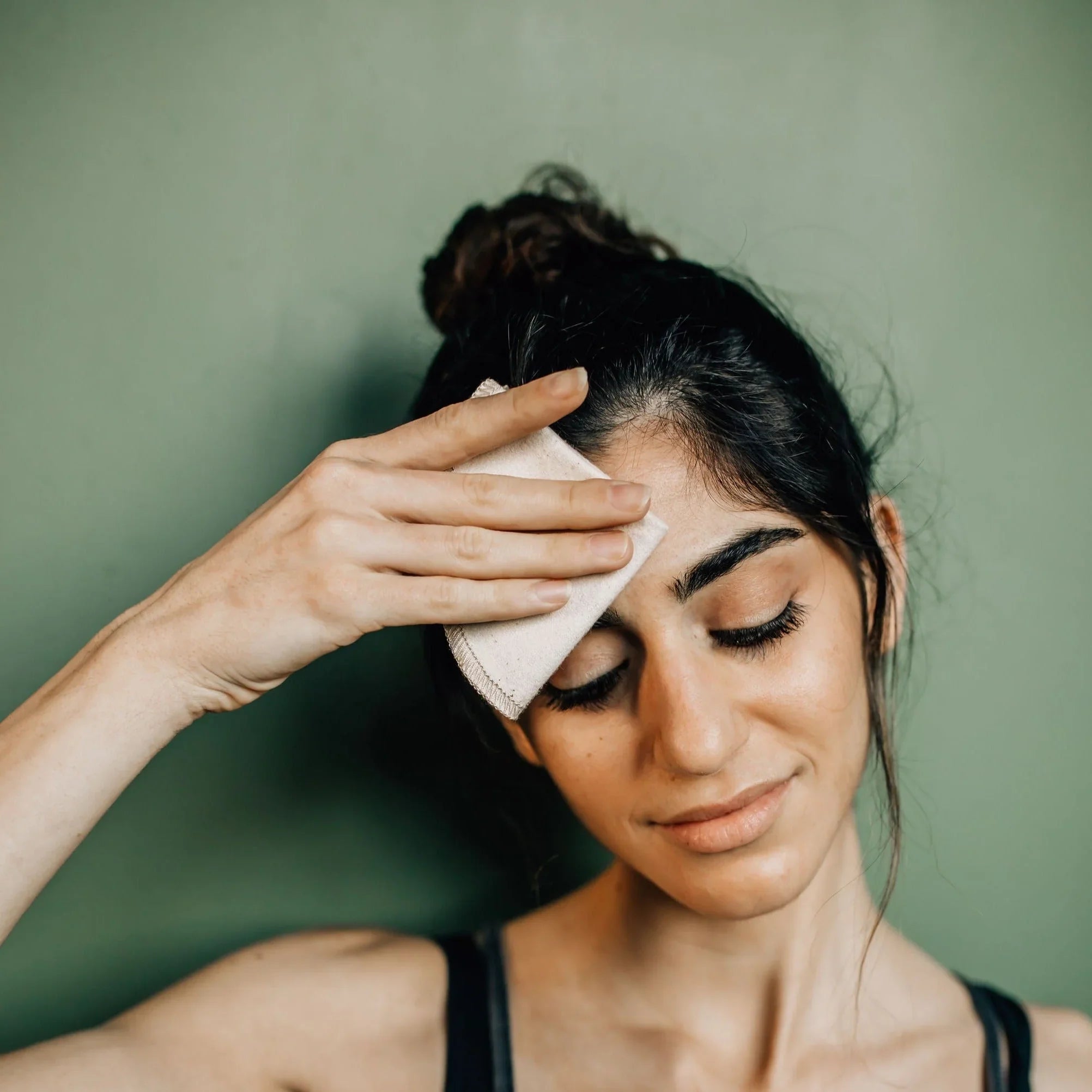 Young woman with dark hair wiping forehead with white cloth against green wall