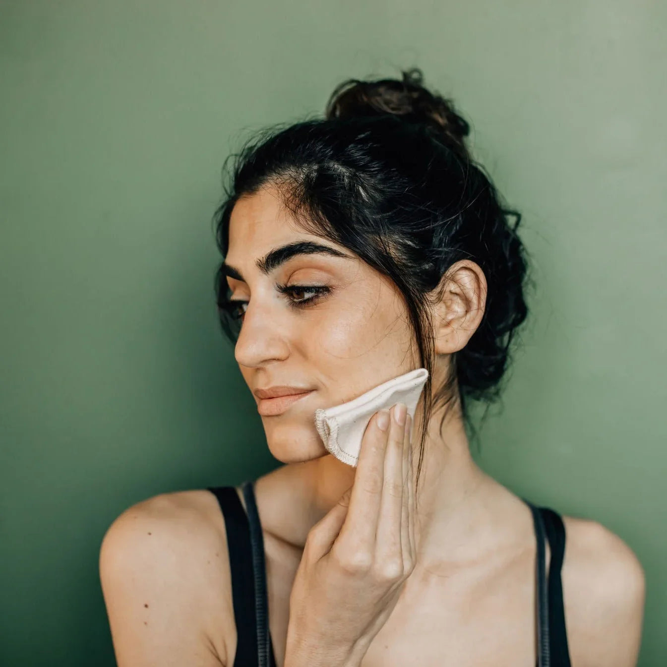 Woman with dark hair in a bun cleansing face with reusable cotton pad against green background