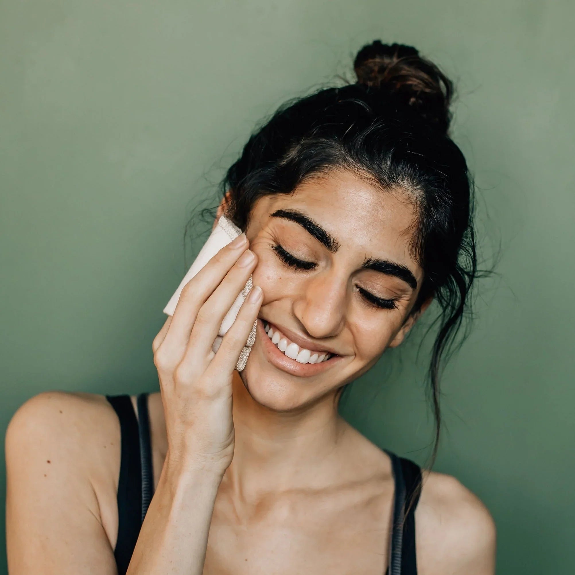 Smiling woman with dark hair in a bun cleansing her face with a cloth against green background