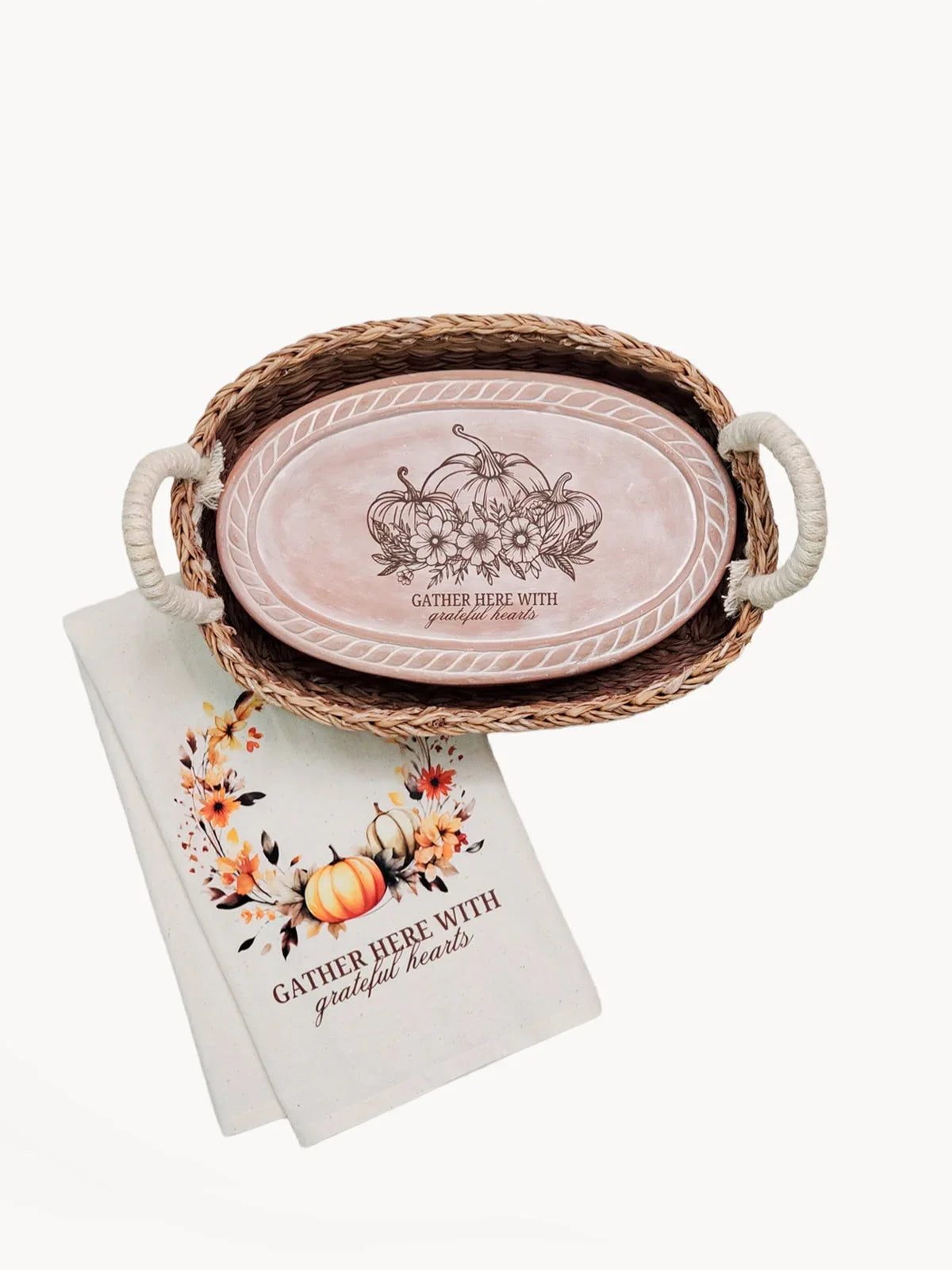 Woven basket with rope handles holding an oval wooden plaque and a floral pumpkin autumn towel both saying Gather Here With Grateful Hearts