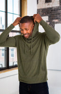 Man wearing olive green hoodie with kangaroo pocket, modeling in natural light by window at Qu!rk Boutique