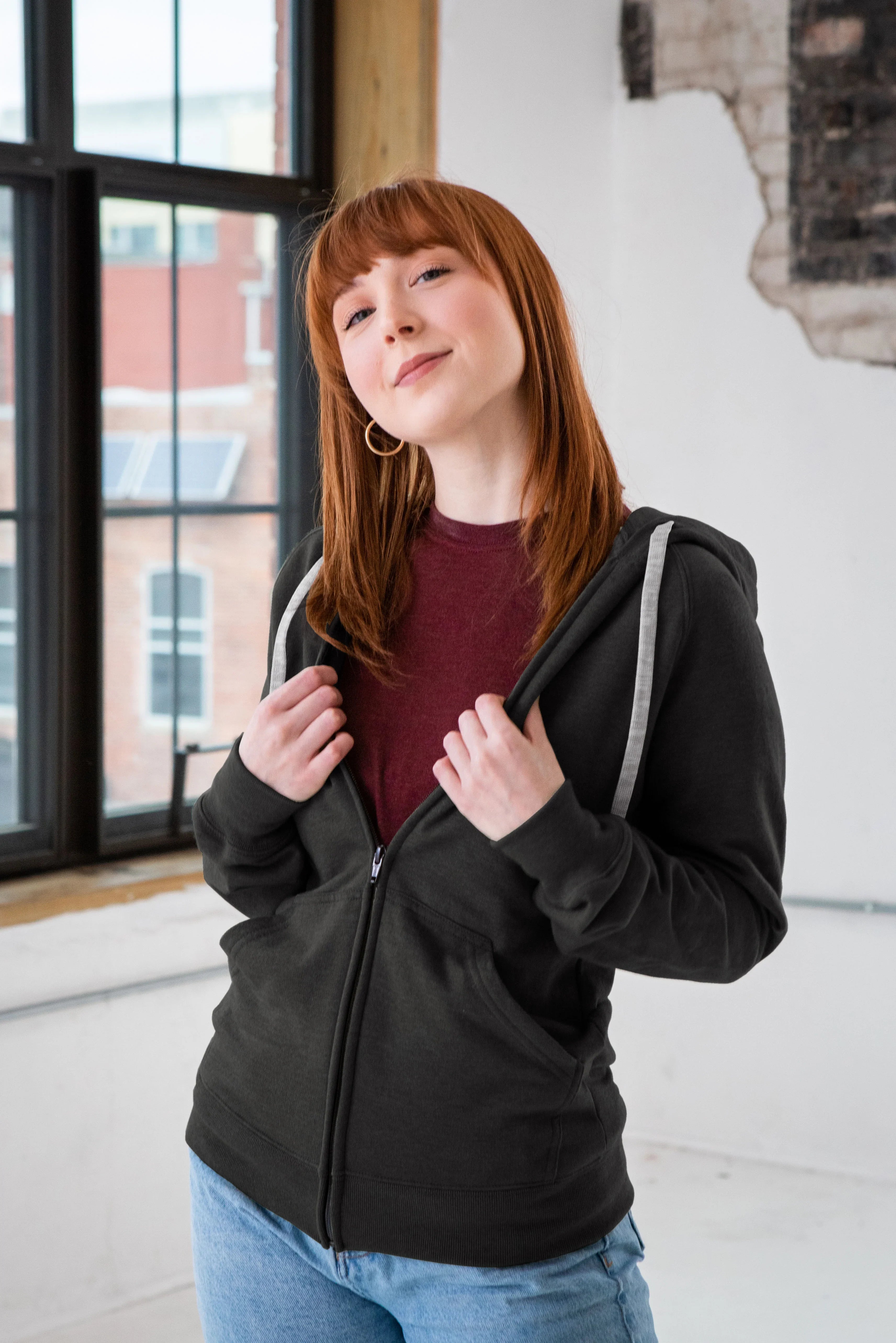 Woman in charcoal zip hoodie and maroon shirt by window, Qu!rk Boutique style