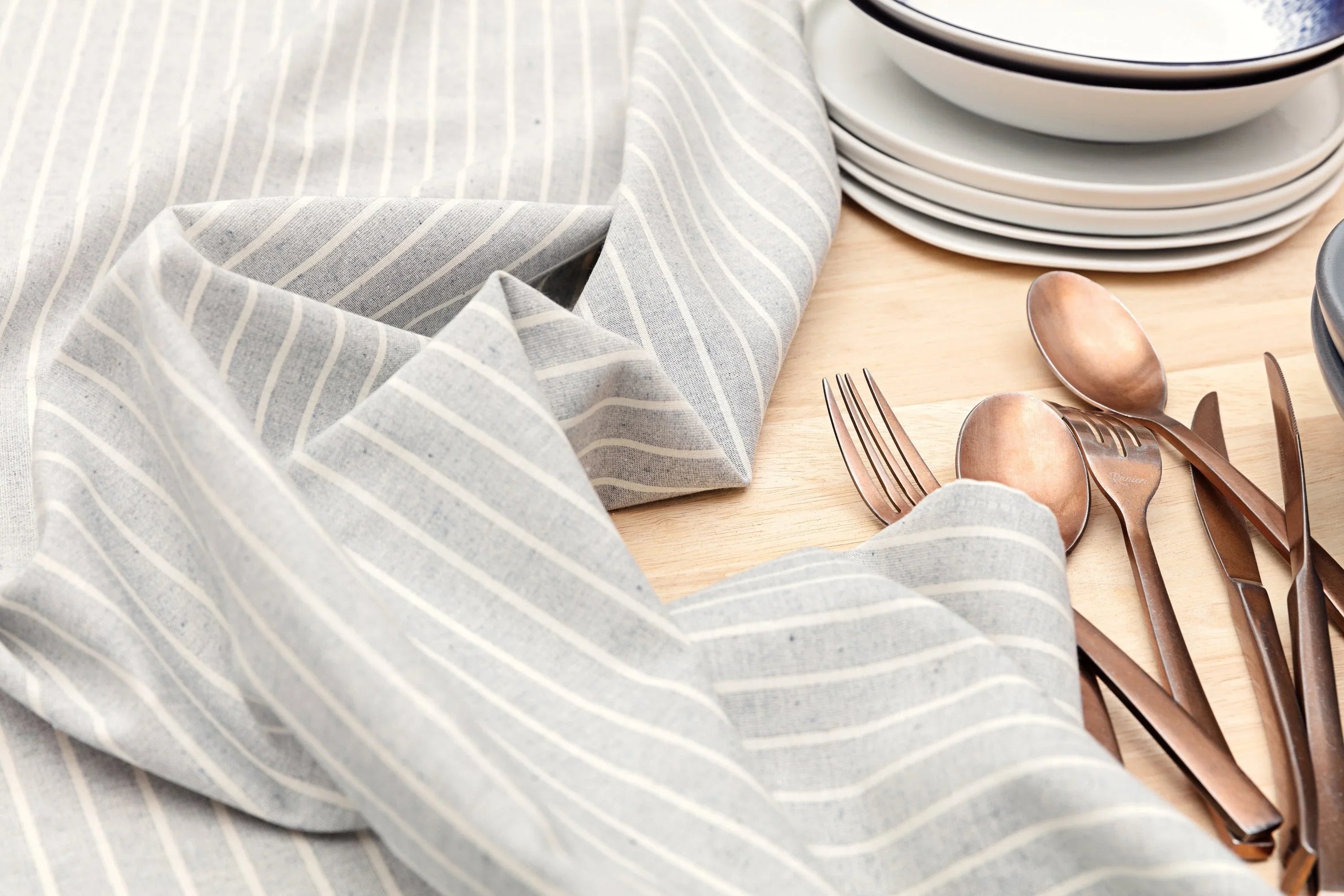 Striped linen napkin, bronze cutlery, and stacked ceramic plates on light wood table
