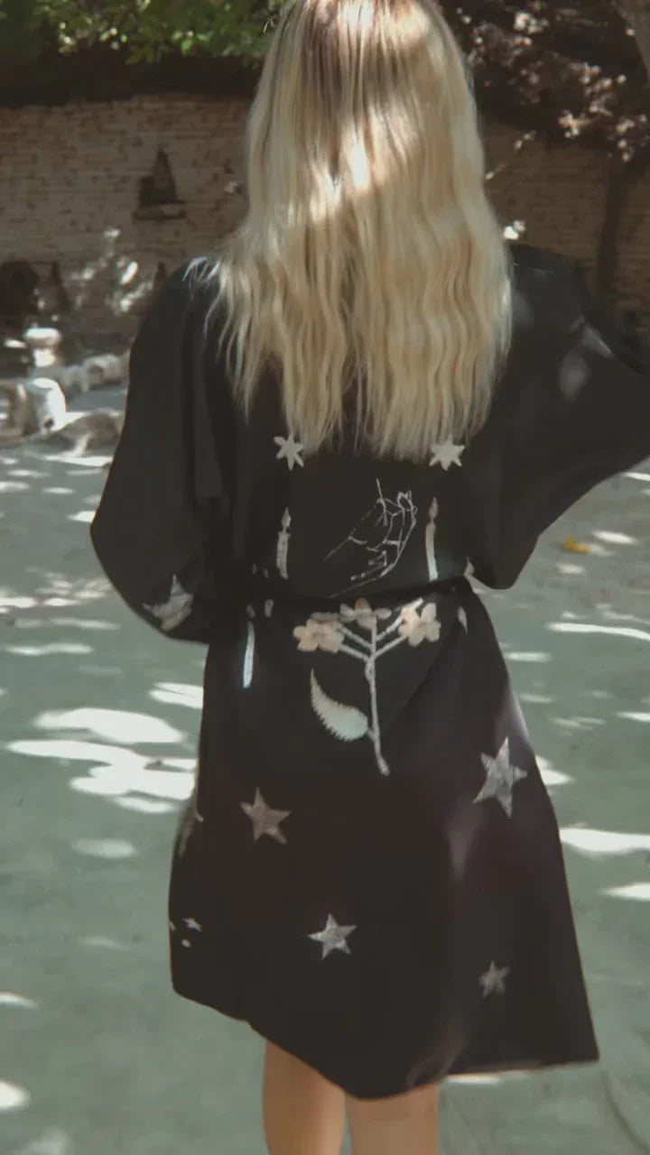 Woman with long blonde hair wearing black robe dress with embroidered stars and floral designs outdoors