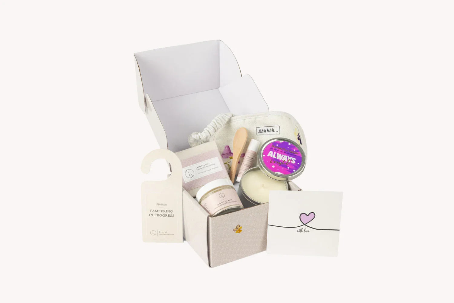 Open gift box with spa essentials including lavender soap, facial mask, candle, lip balm, wooden spoon, and pampering door hanger