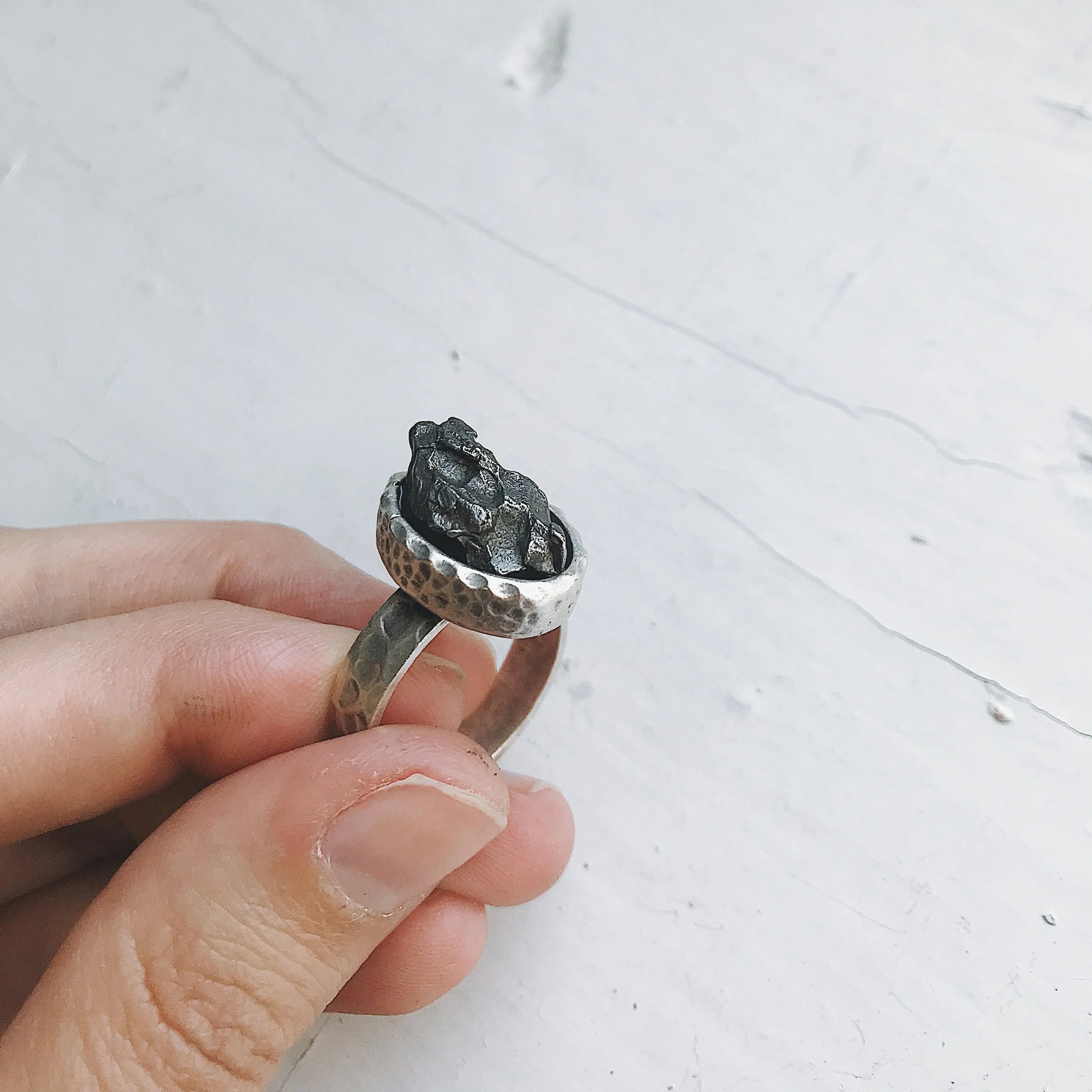 Hand holding a silver hammered ring featuring a textured, dark metallic stone on a light gray background