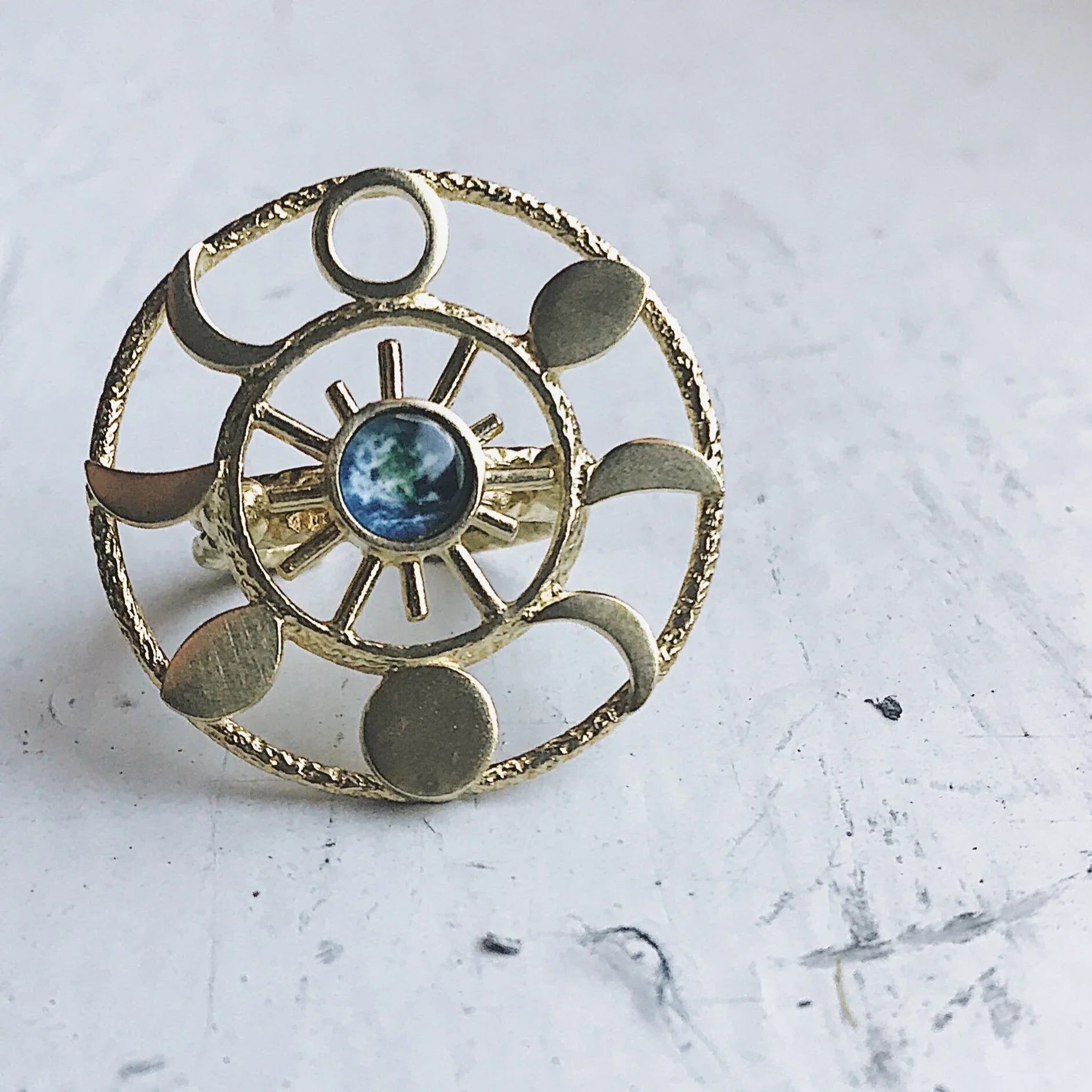 Gold celestial ring with sun, moon phases, and Earth centerpiece on textured white surface