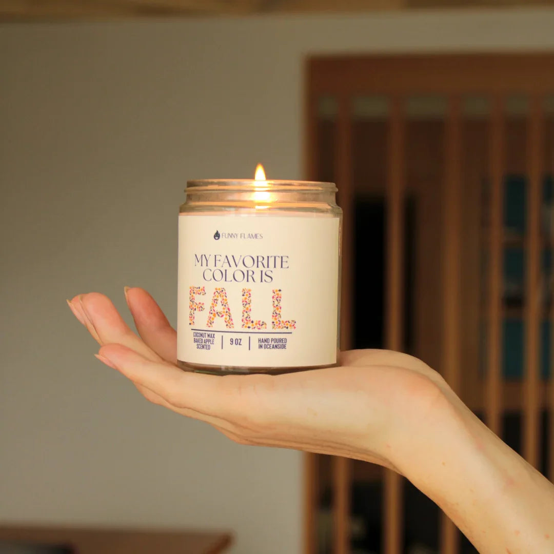 Hand holding a lit scented candle labeled 'My Favorite Color Is Fall' with floral print text