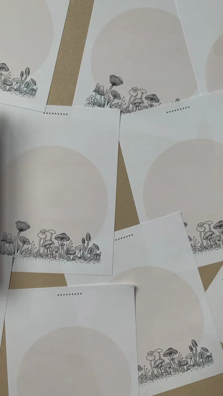 Close-up of white cards with beige circles and black line art of mushrooms and plants on a brown surface