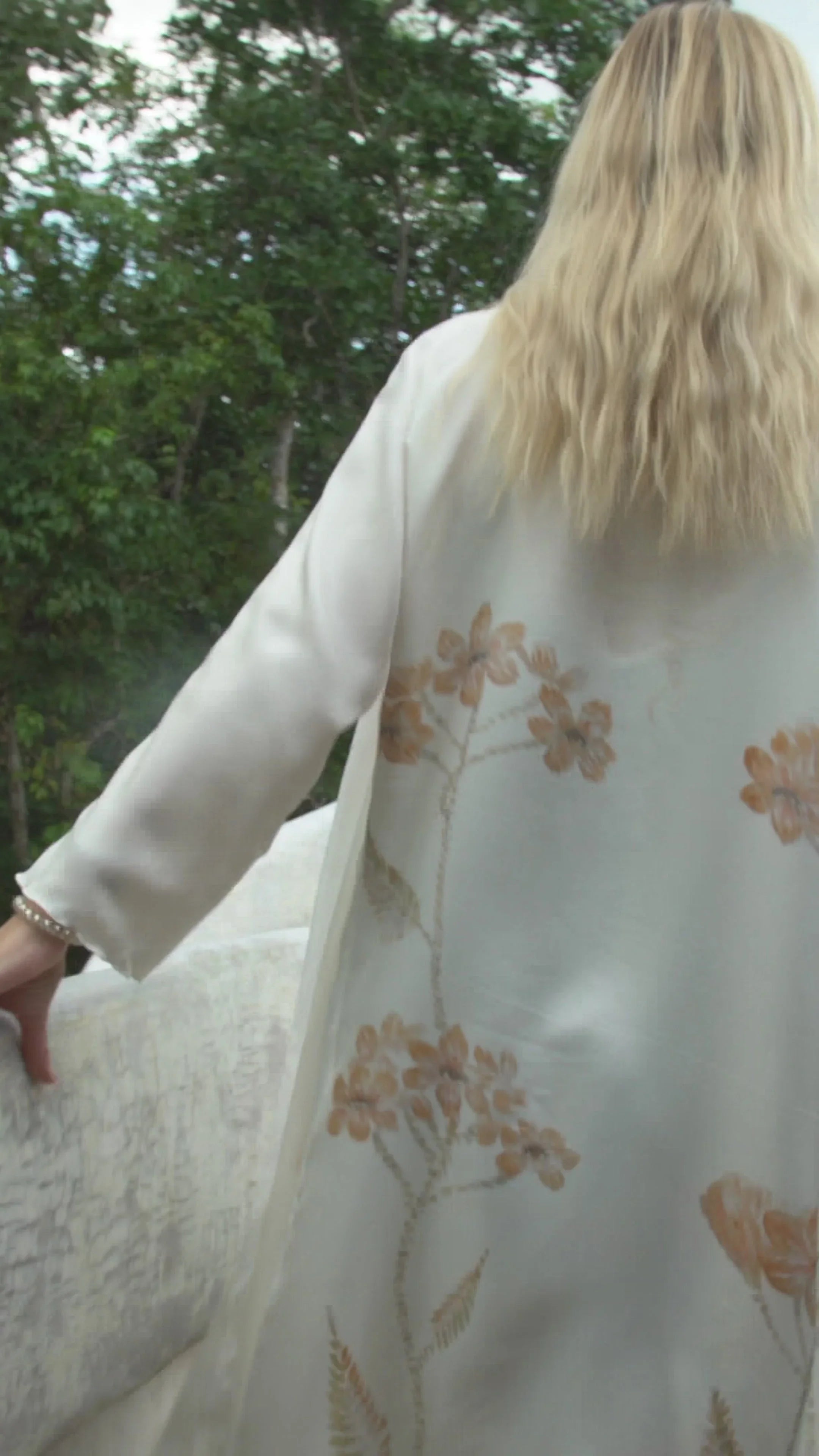 Woman with blonde wavy hair wearing white floral embroidered cardigan outdoors by white textured railing