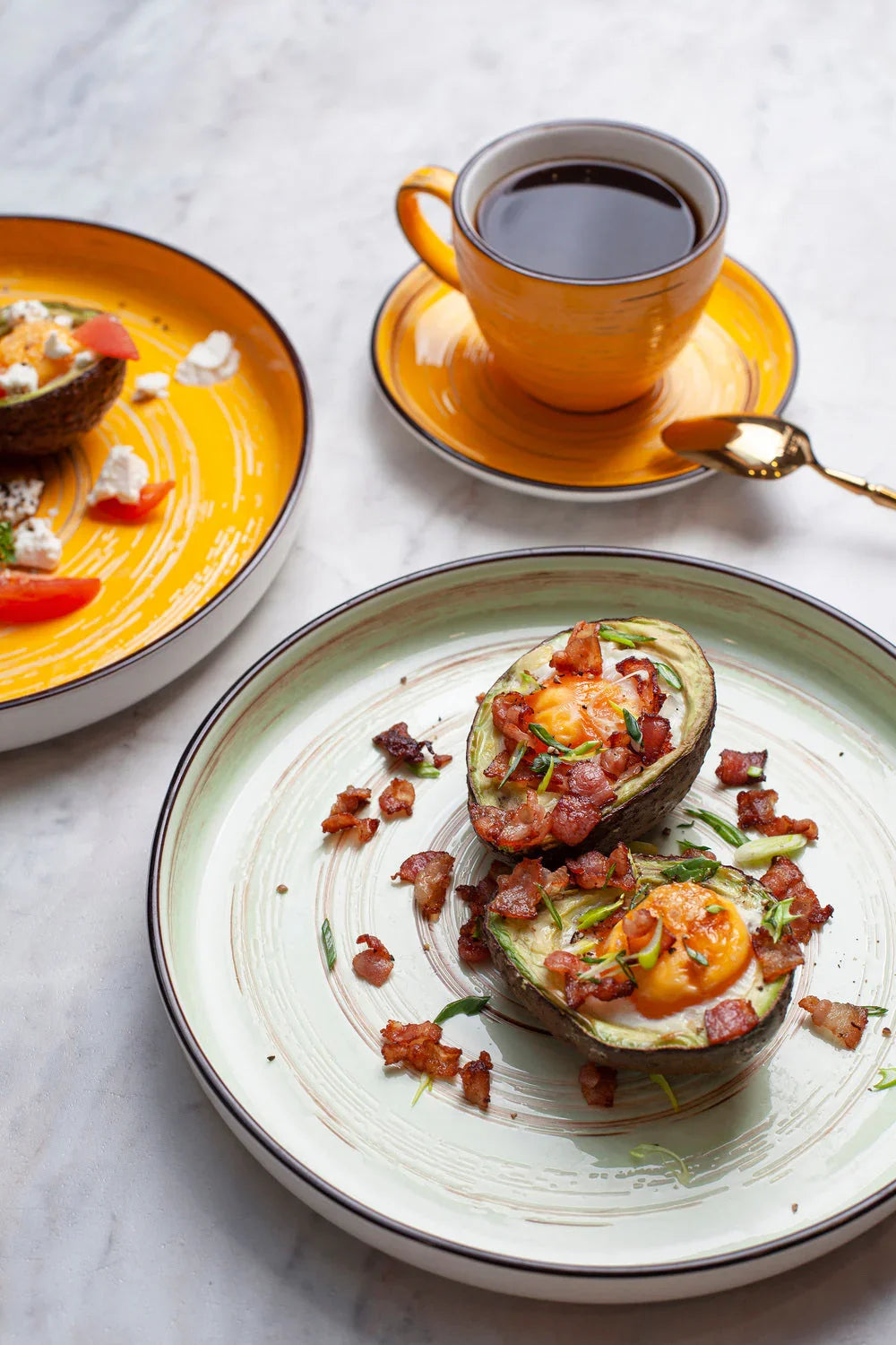 Brunch scene with baked avocado halves topped with bacon and eggs, coffee in orange cup, and colorful plates.